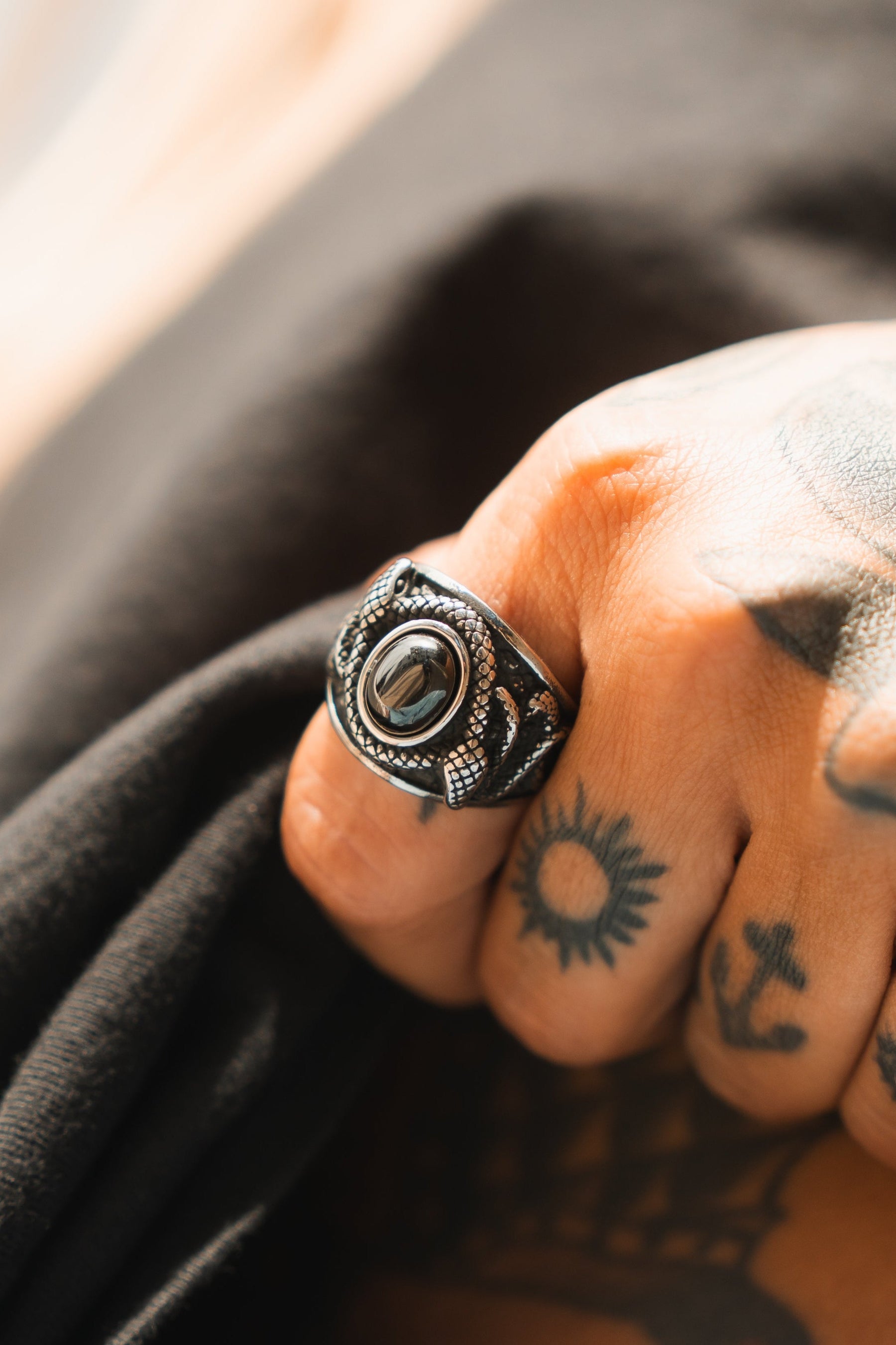 A tattooed hand, featuring a sun and an anchor, showcases the handcrafted Relentless Betrayal Onyx Rebirth Ring with its glossy onyx stone and intricate design. The stainless steel band harmonizes elegantly with the persons dark clothing.