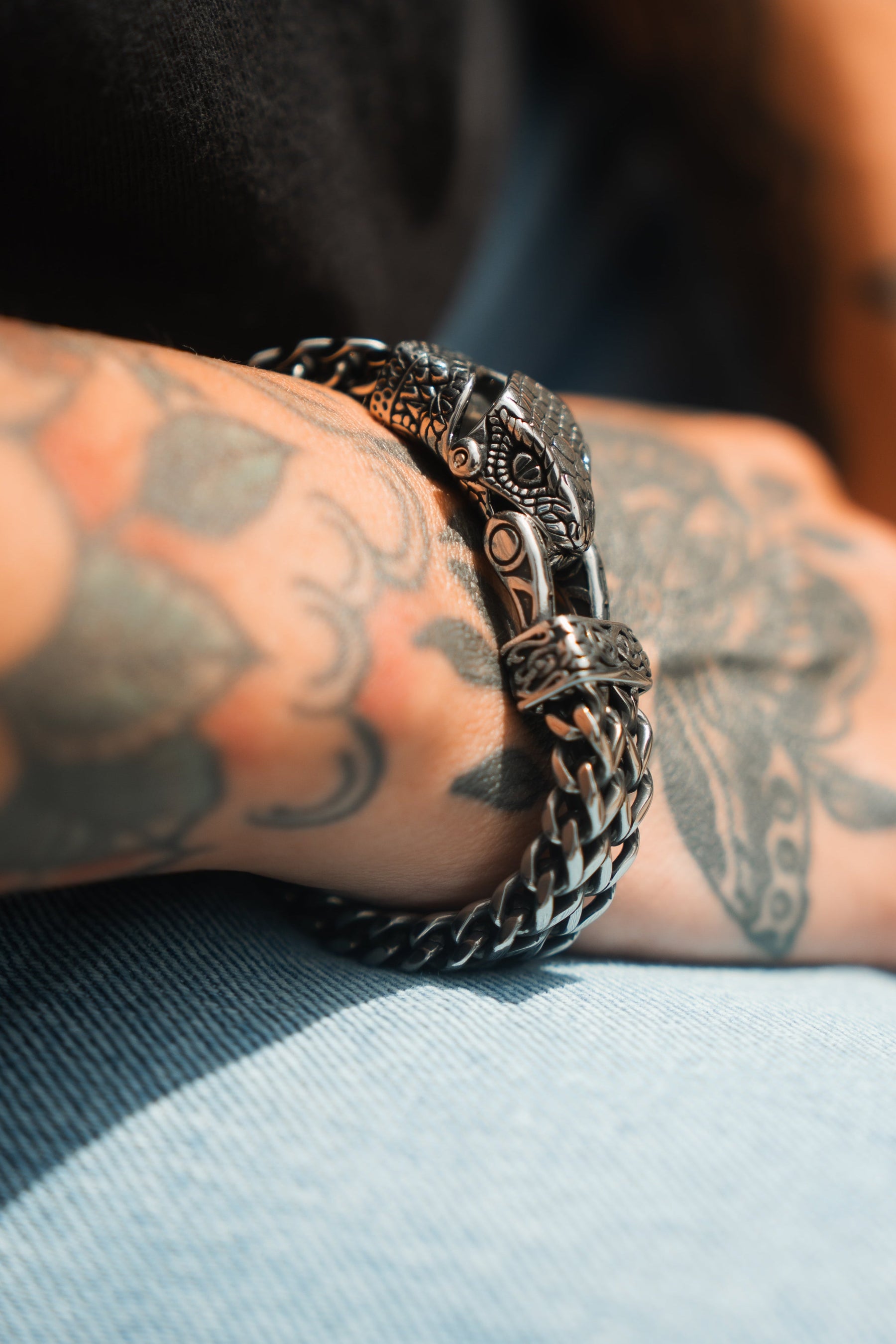 A stainless steel Viper Bracelet by Relentless Betrayal adorns a tattooed arm resting on denim. Intricate floral and abstract tattoos complement the bracelets design, highlighted under bright lighting.