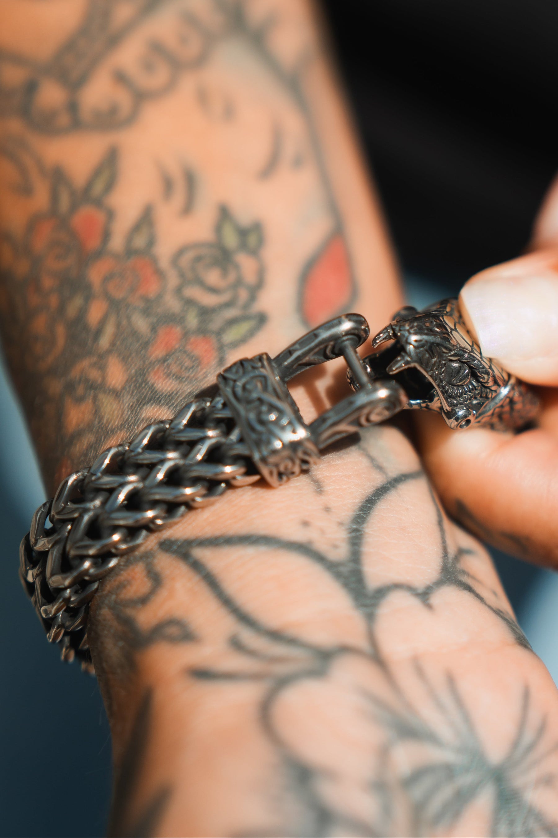 Close-up of a tattooed arm highlights a detailed Relentless Betrayal Viper Bracelet with an intricate clasp, complementing floral tattoos in color and black ink. The person grasps the stainless steel bracelet, adding elegance to their style.