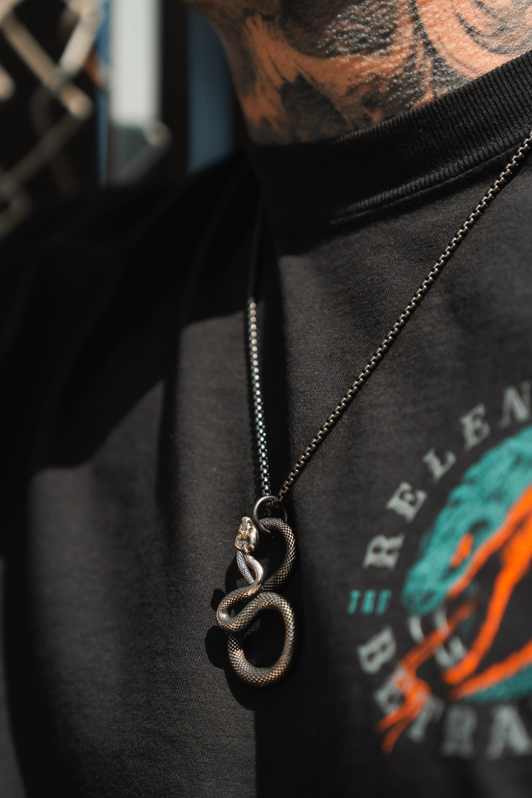 Close-up of a person wearing a black shirt with colorful graphics, showcasing serpentine elegance with the Slither Necklace by Relentless Betrayal. The background is slightly blurred, revealing neck tattoos, as the statement piece features an intricate snake pendant.