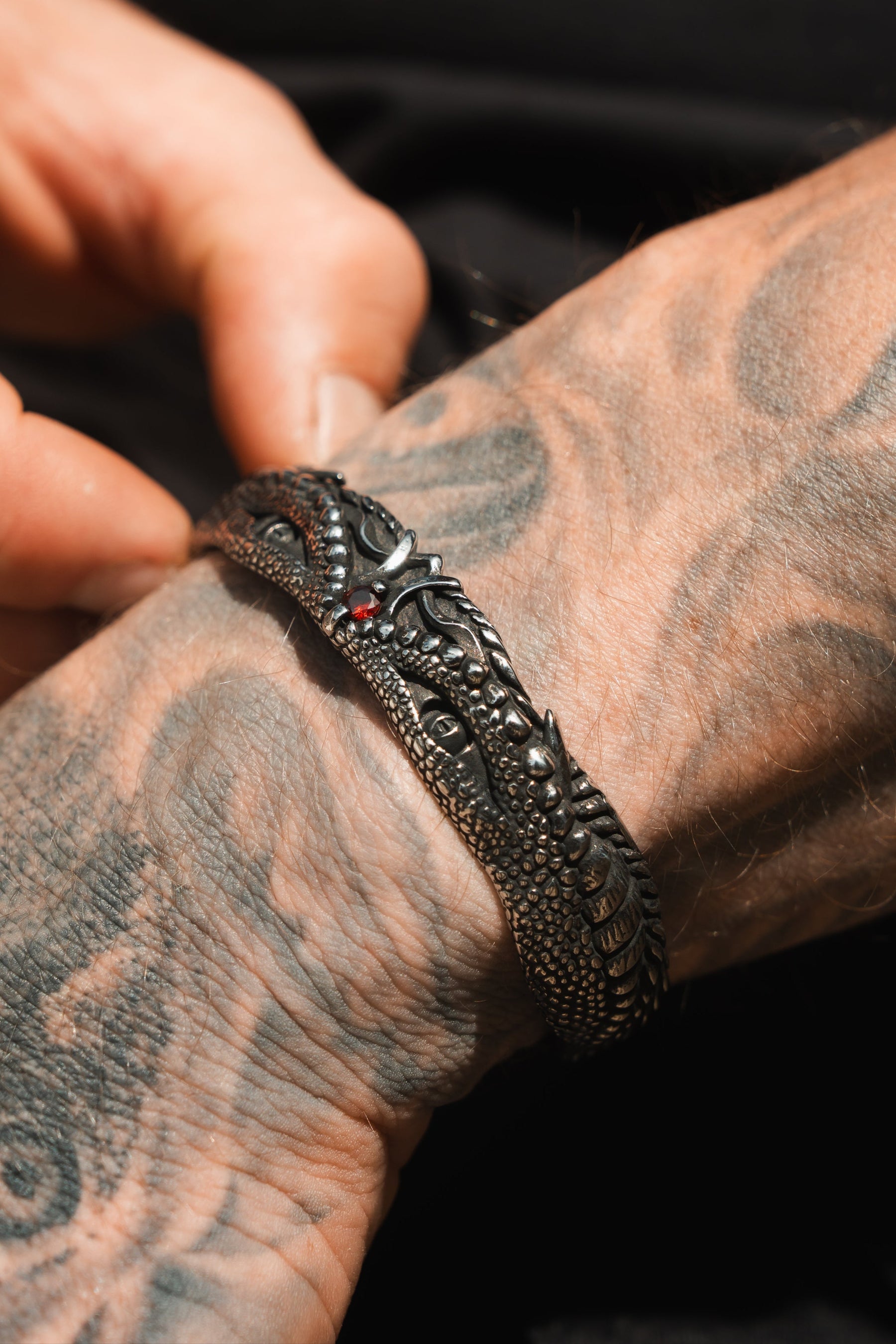 A tattooed hand adjusts Relentless Betrayals Holy Grail Bracelet, a stainless steel masterpiece with intricate designs and red accents, while abstract patterns decorate the skin.