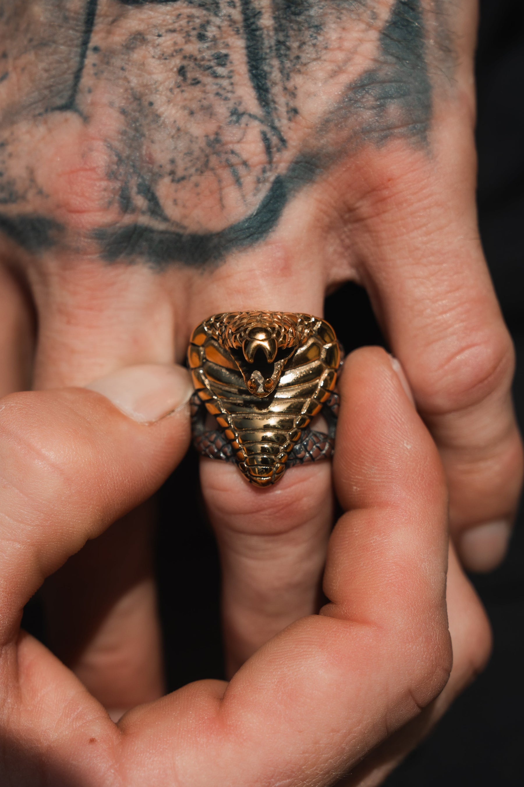 A close-up shows a hand with the handcrafted Cobra King Ring by Relentless Betrayal on the ring finger, while the other hand adjusts it. Abstract tattoos adorn the skin, adding to this striking pieces unique allure.