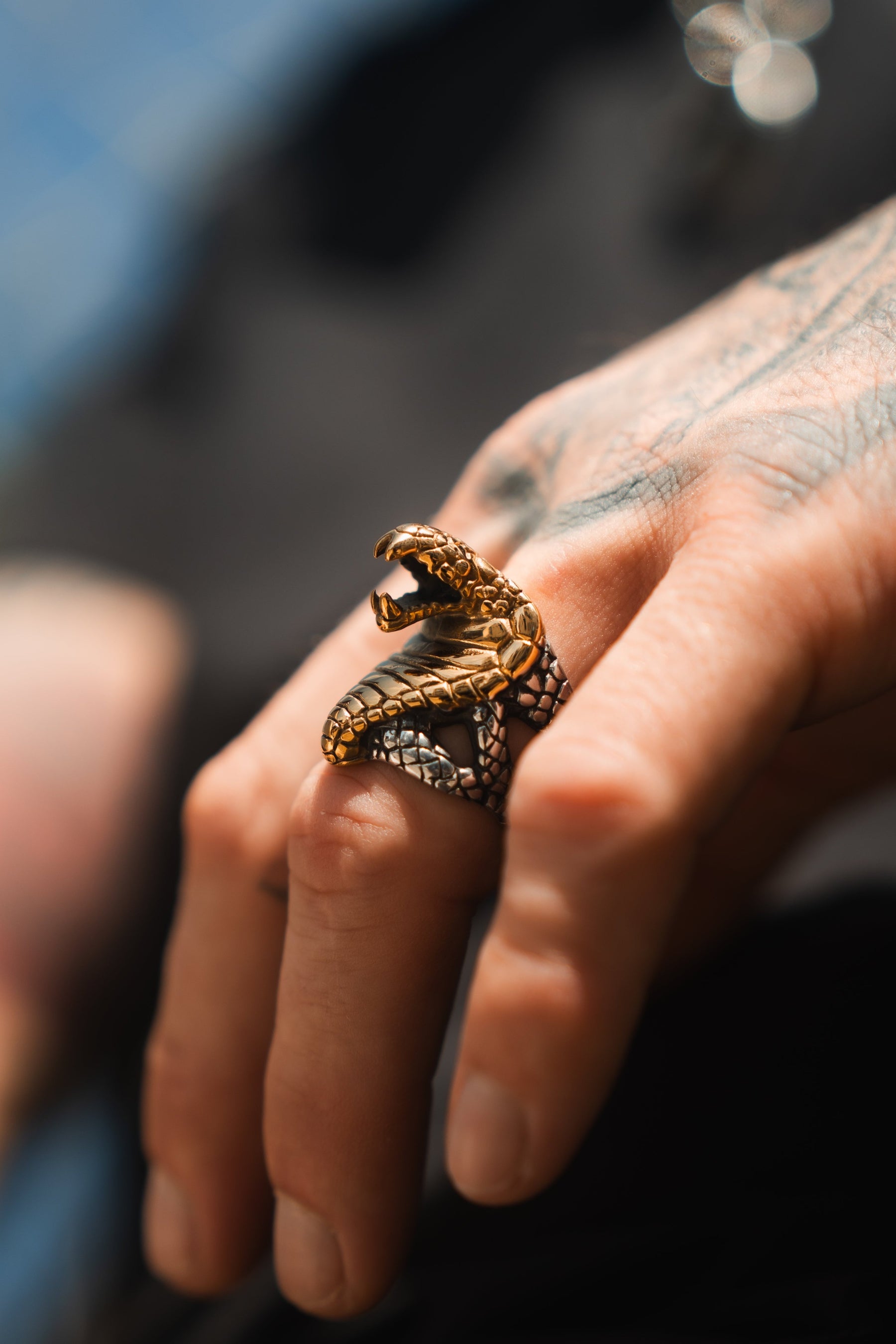 A close-up showcases a hand wearing the Cobra King Ring by Relentless Betrayal, shaped like a birds talon gripping the finger. Its intricate scale and claw design stand out against a blurred background highlighting part of a tattoo.