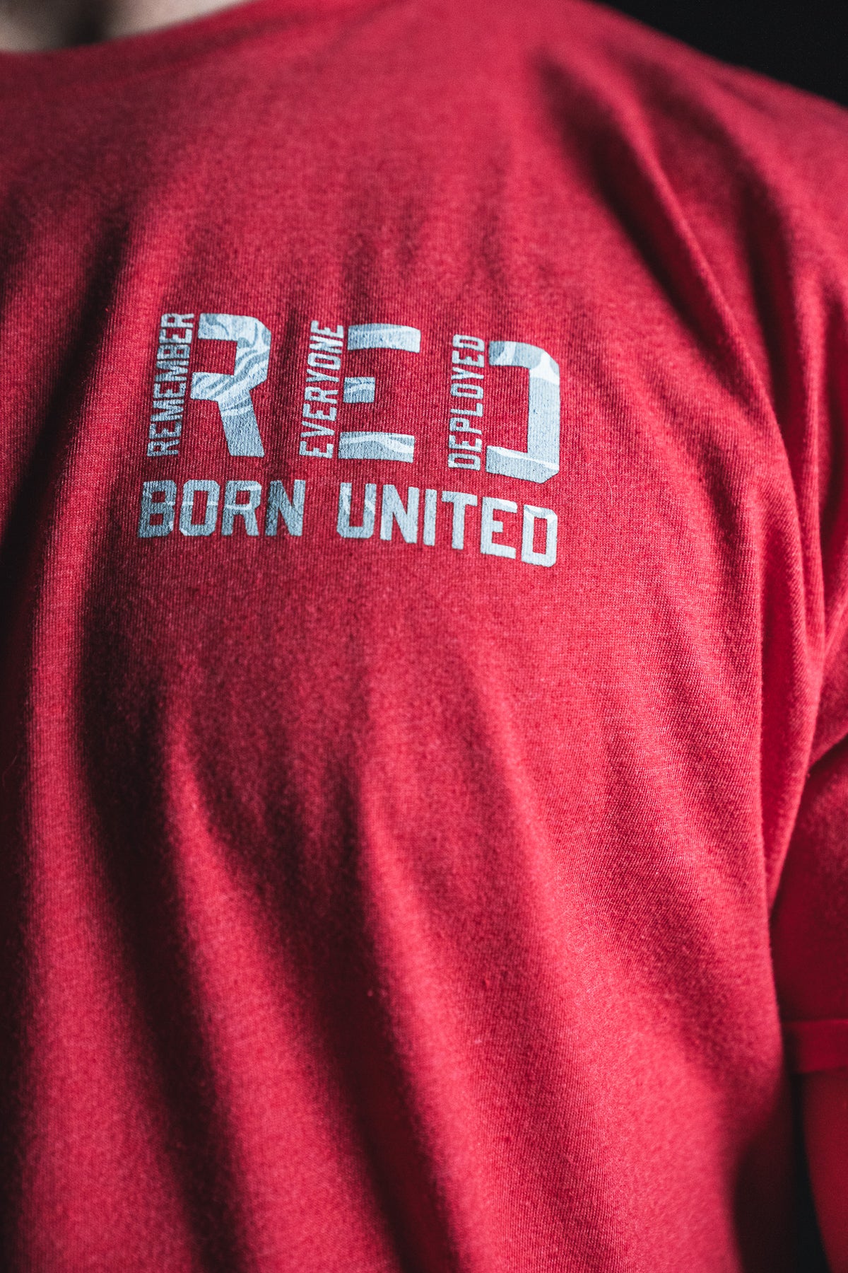 A person wearing a Born United Private Label RFV2 red T-shirt with “REMEMBER EVERYONE DEPLOYED RED BORN UNITED” printed in white on the chest.