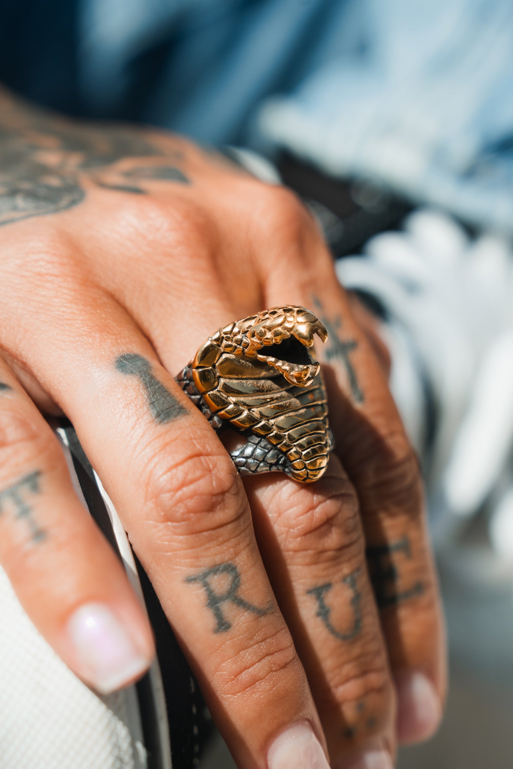 A close-up reveals a tattooed hand wearing the handcrafted Cobra King Ring by Relentless Betrayal. This intricately detailed stainless steel piece features a snake head with textured scales, showcasing the artistry of both jewelry and tattoos.