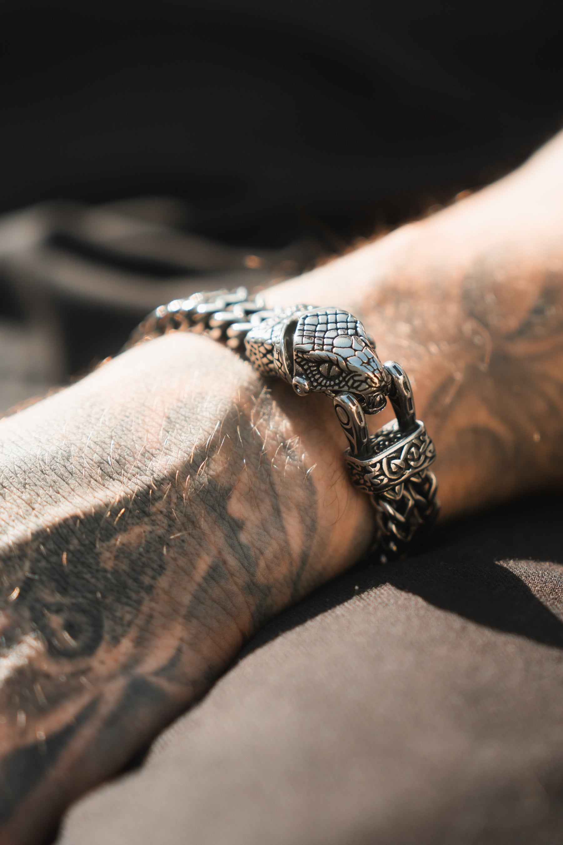 Close-up of a tattooed forearm wearing the Relentless Betrayal Viper Bracelet, highlighting its intricate snake head design. This stainless steel piece features textured links, with the softly blurred background enhancing its exquisite craftsmanship.