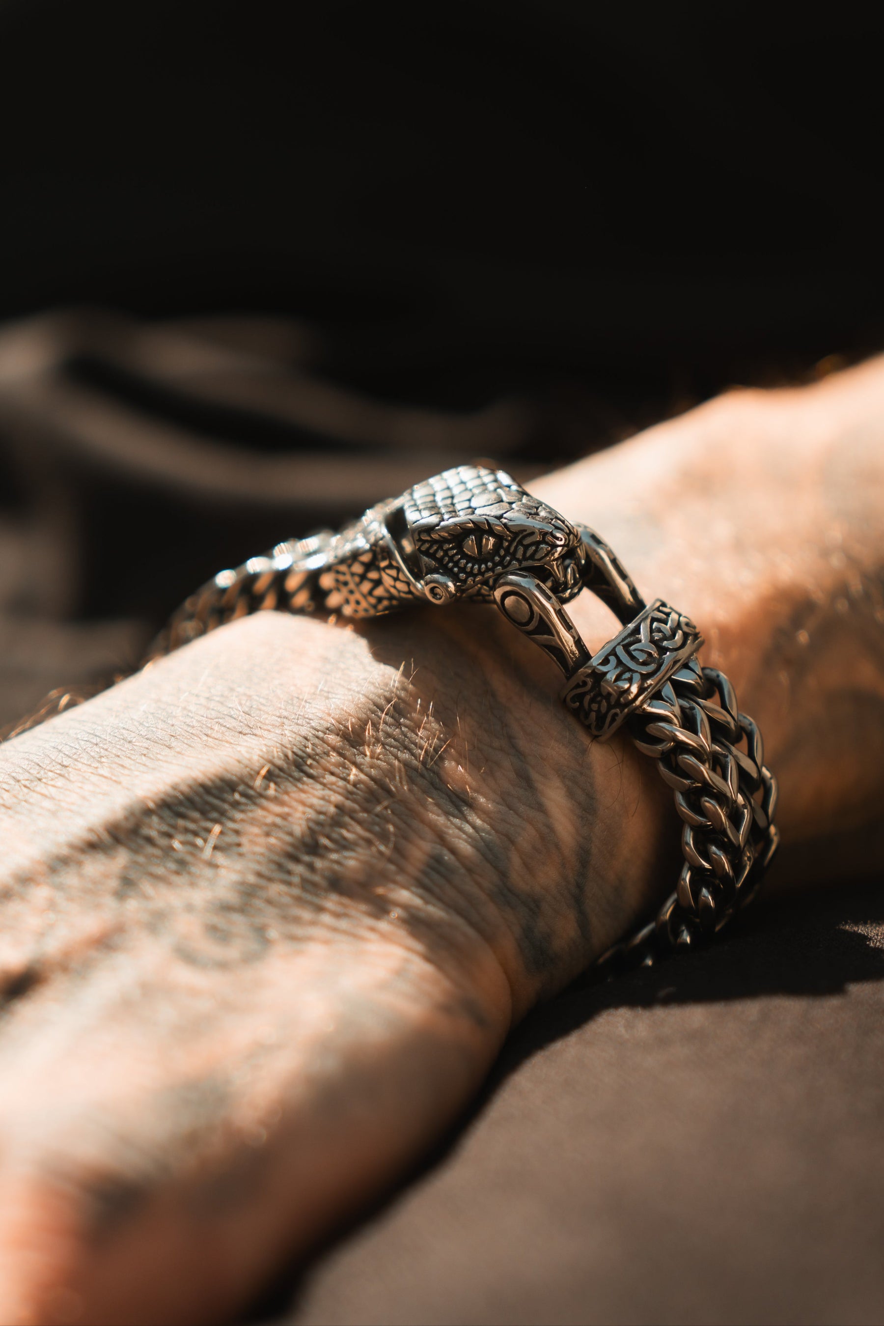 A tattooed hand sports the stainless steel Viper Bracelet by Relentless Betrayal, featuring intricate scales and snake head designs. The softly blurred background highlights the detailed craftsmanship of both the bracelet and the hand.