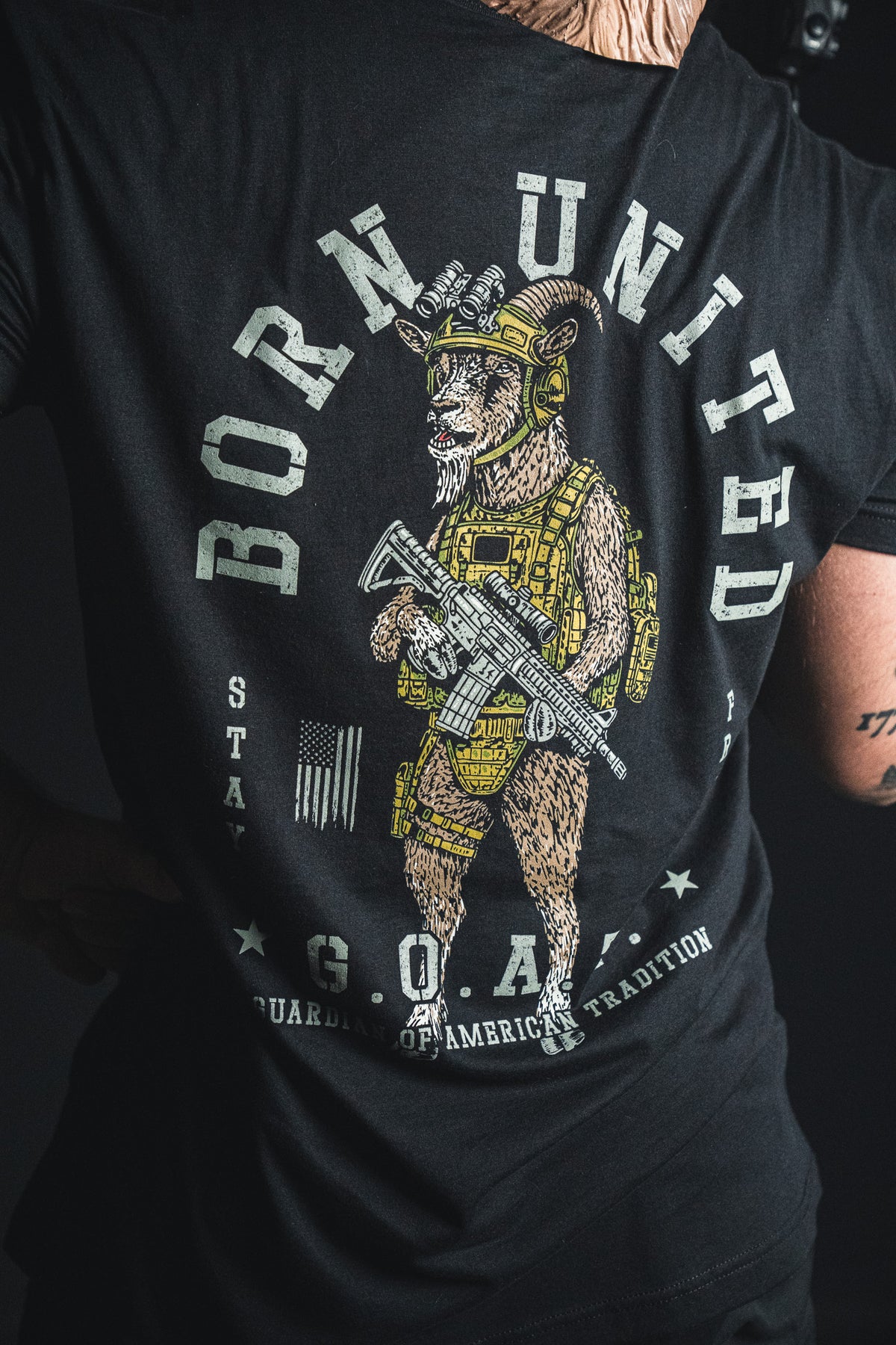 A person sports the G.O.A.T. shirt from Born United Private Label—a black tee showcasing an illustrated goat in military gear with a rifle, featuring “BORN UNITED” and “G.O.A.T.” on the back for a bold display of American tradition and patriotic pride.
