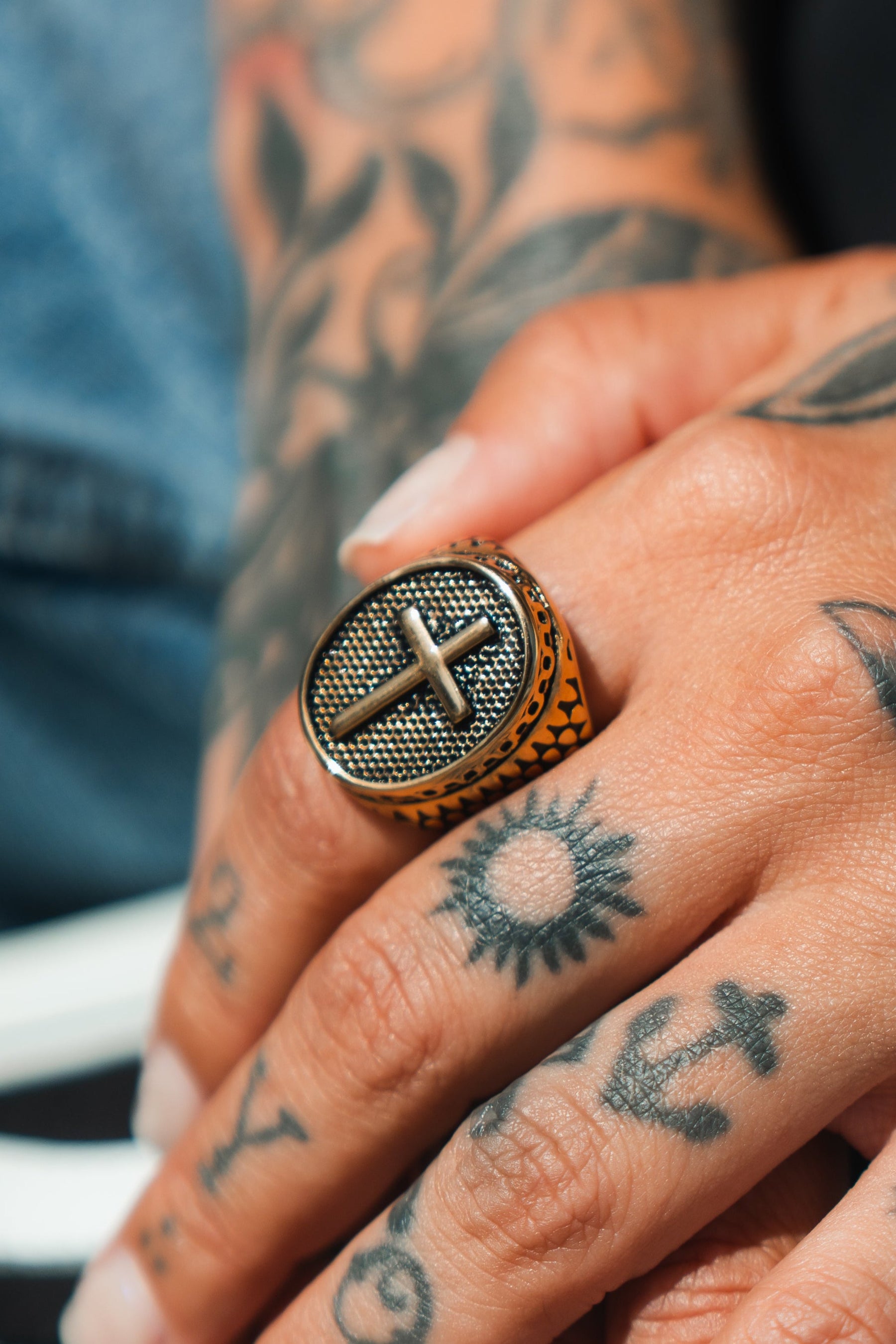 Close-up of a tattooed hand with a sun and anchor, wearing an impressive Risen Savior Ring- Gold by Relentless Betrayal, handcrafted in stainless steel with a bold cross design.