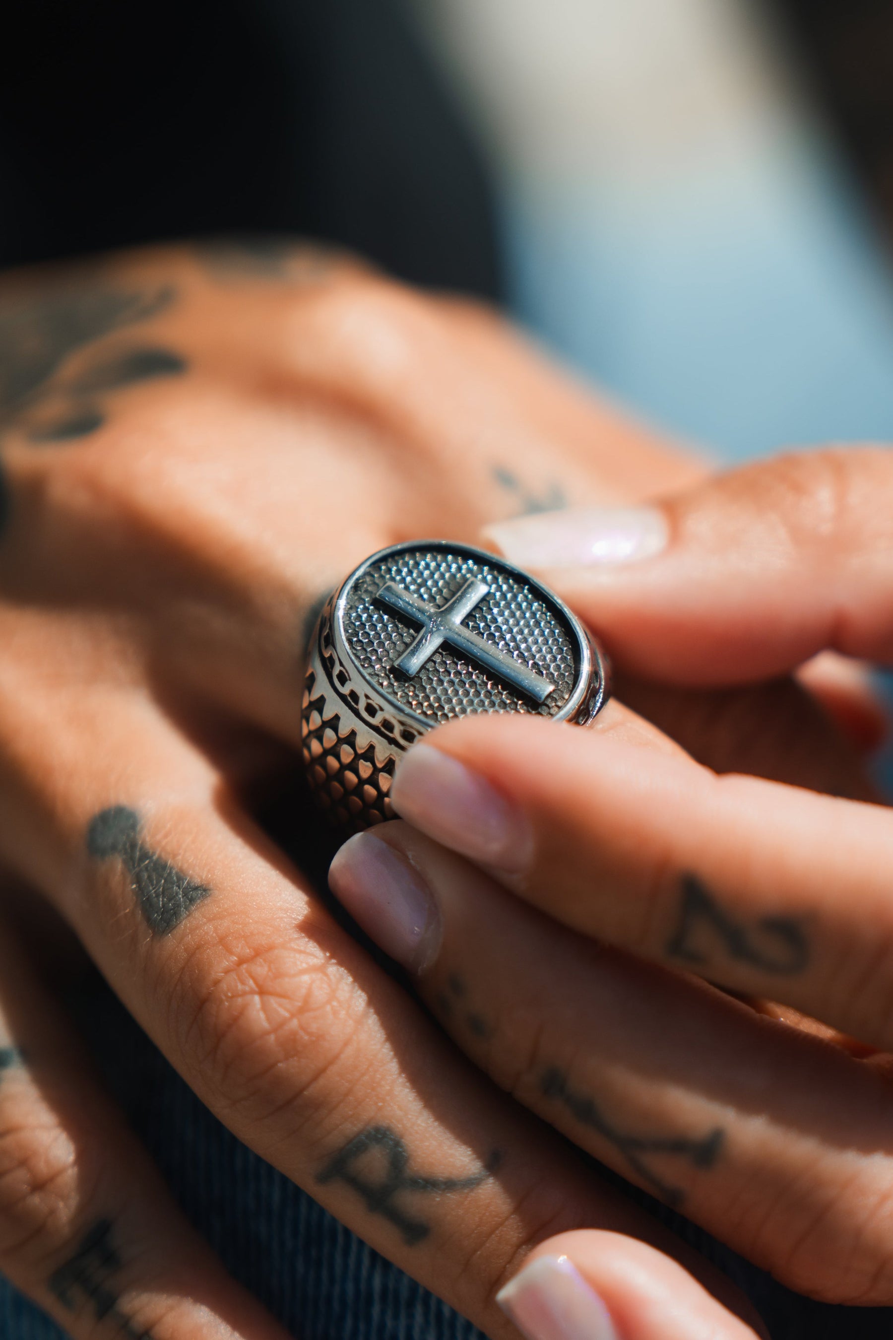 A tattooed hand holds the Risen Savior Ring-Silver by Relentless Betrayal, showcasing a raised, textured cross design. Handcrafted from stainless steel, the ring features intricate patterns along the band. Neat nails and natural light accentuate both the tattoos and this exquisite piece.