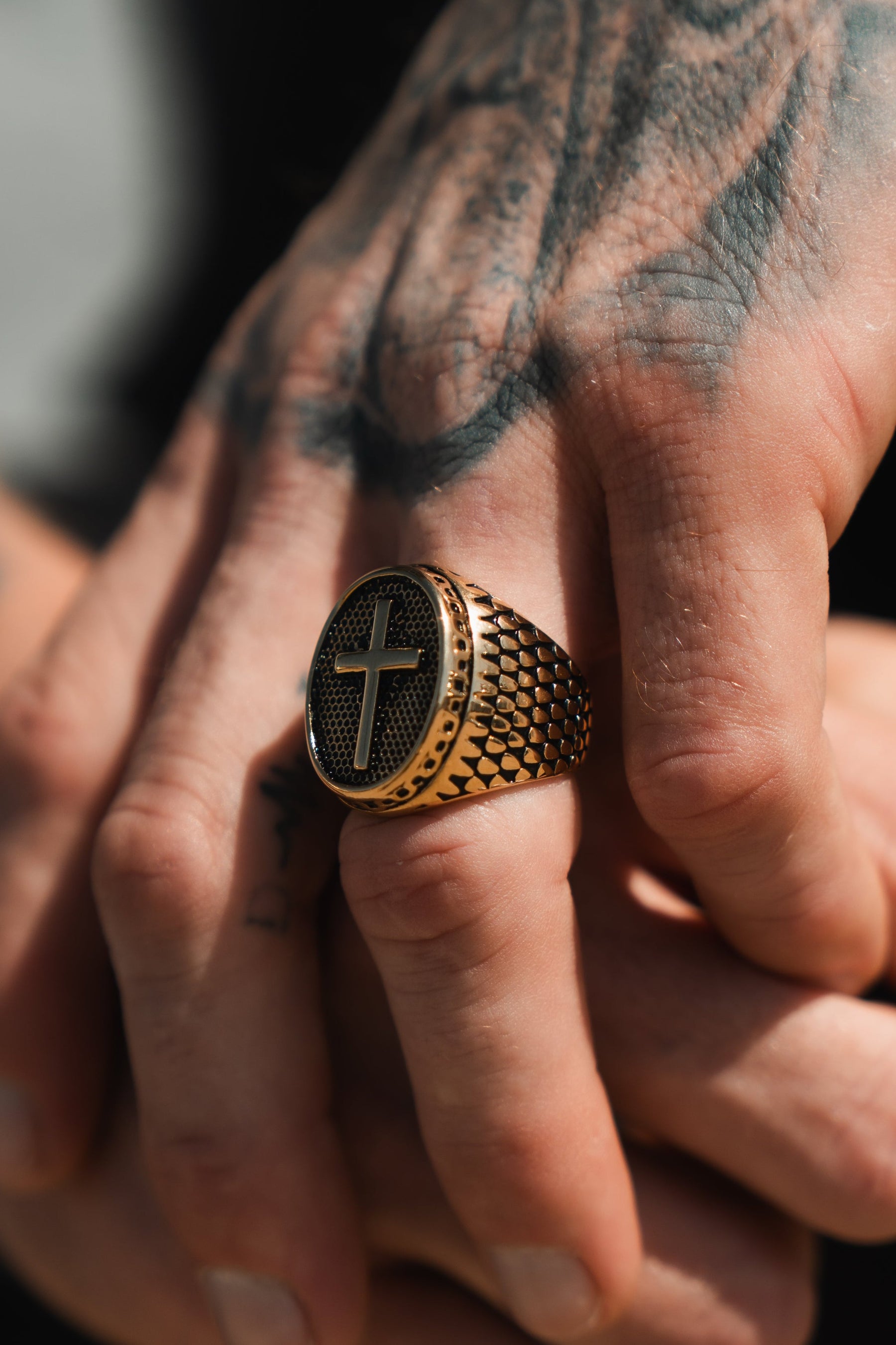 Tattooed hands feature the Risen Savior Ring- Gold by Relentless Betrayal, a textured gold piece with a cross design. The tattoos and stainless steel enhance its handcrafted details, while lighting highlights its intricate beauty.
