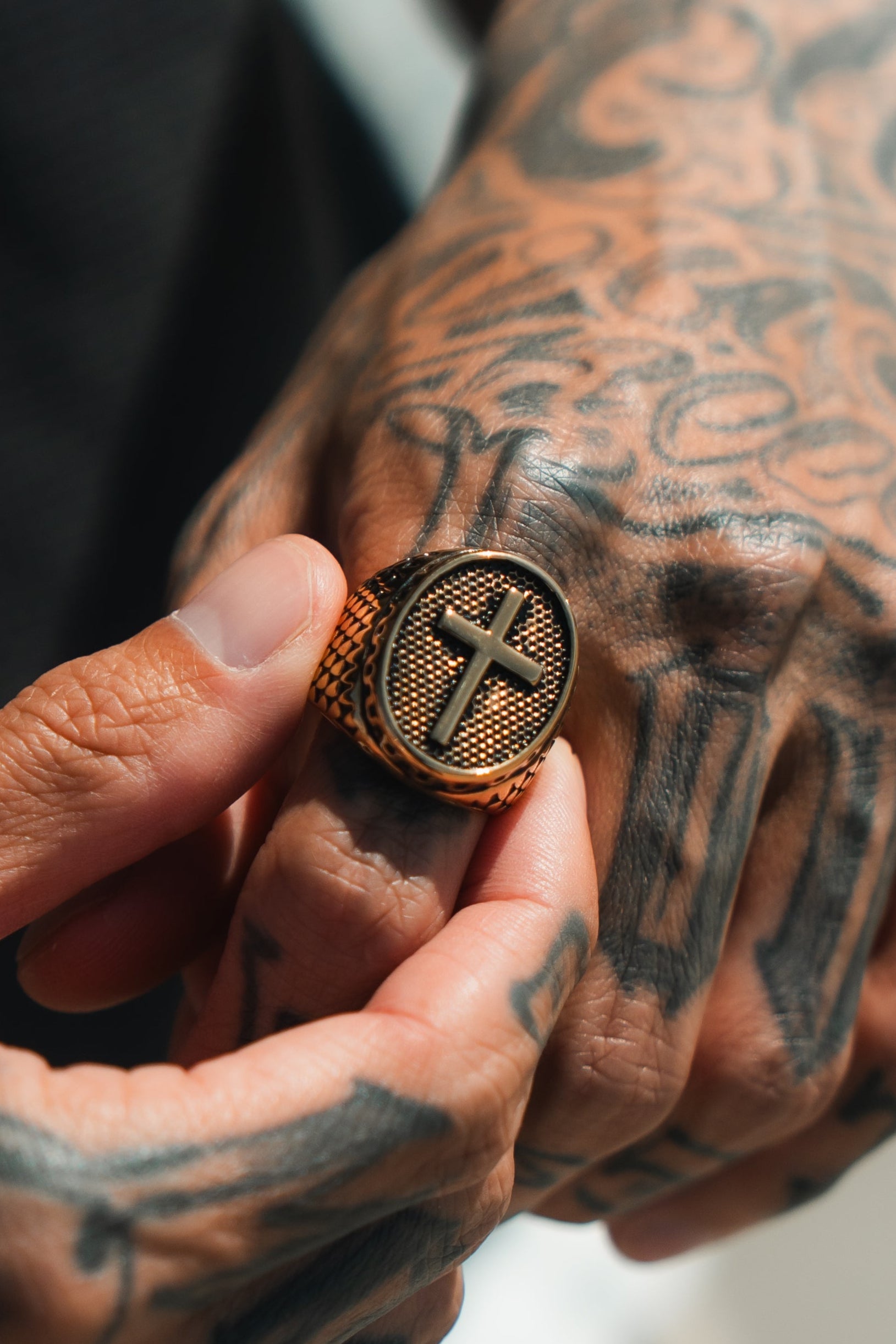 A tattooed hand displays the Risen Savior Ring- Gold by Relentless Betrayal, showcasing a raised cross design on a textured band. The detailed tattoos enhance the rings elegance, while the softly blurred background amplifies its exquisite allure.