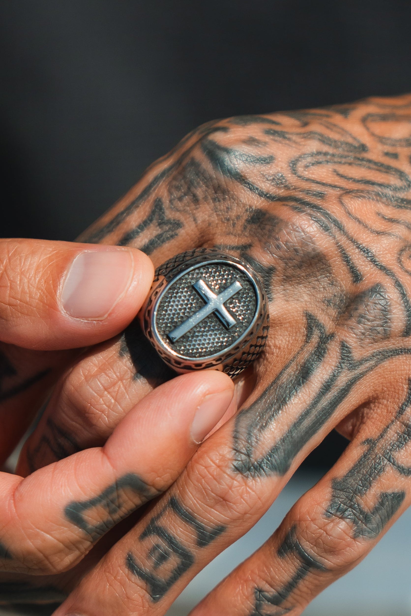 A tattooed hand adorned with Relentless Betrayals handcrafted Risen Savior Ring in silver, featuring a raised cross design, is positioned against a blurred background, accentuating the ring and tattoo details.