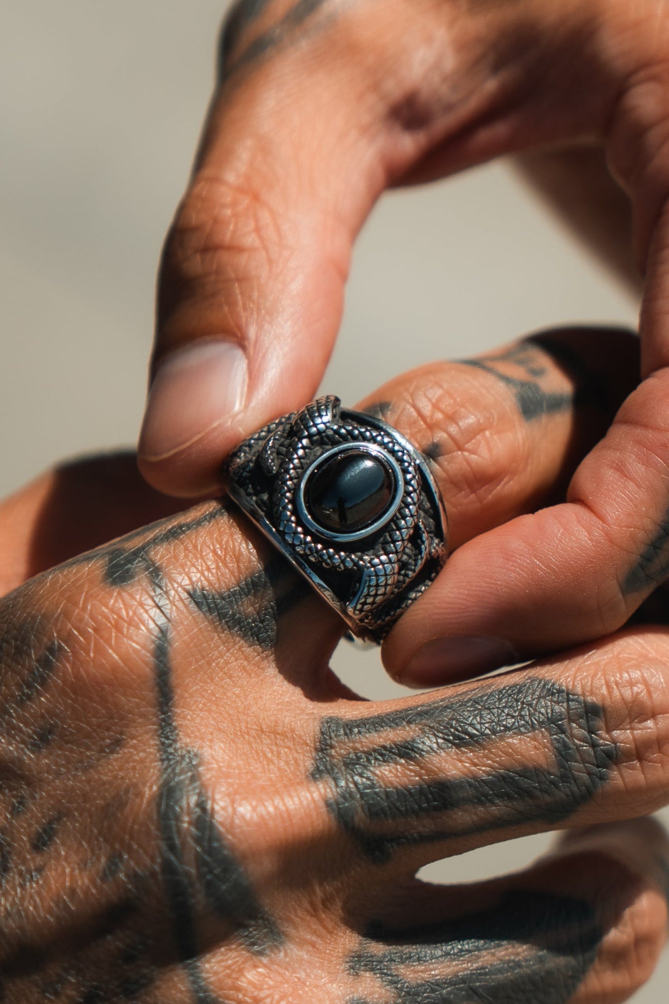 A tattooed hand delicately pinches the Onyx Rebirth Ring by Relentless Betrayal, featuring a textured silver band with an ornate design and round black stone center, against a softly blurred background.