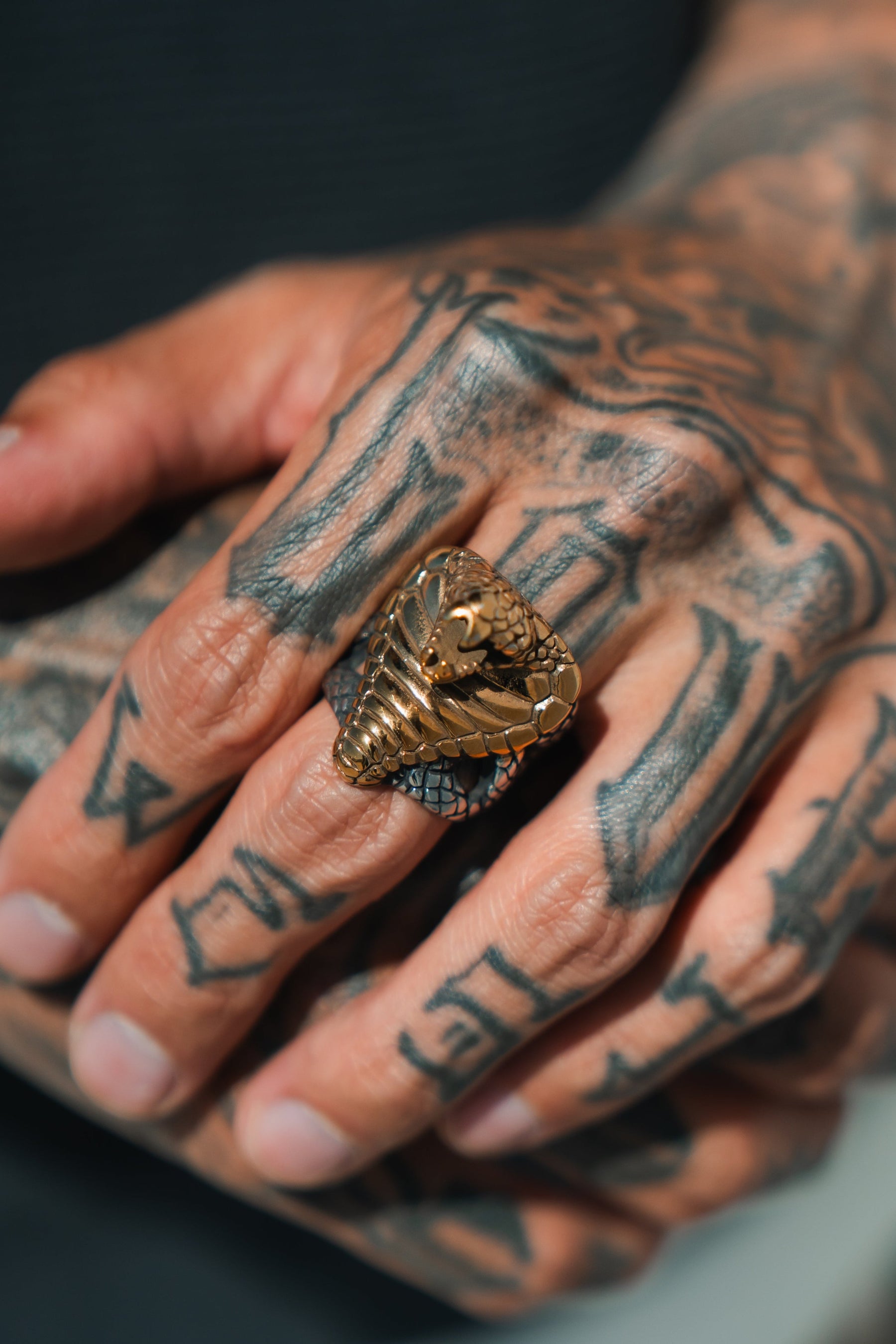 A tattooed hand displays the intricately handcrafted Cobra King Ring by Relentless Betrayal, with detailed scales and a hood that wraps elegantly around the finger, its serpentine beauty enhanced by stainless steel artistry.