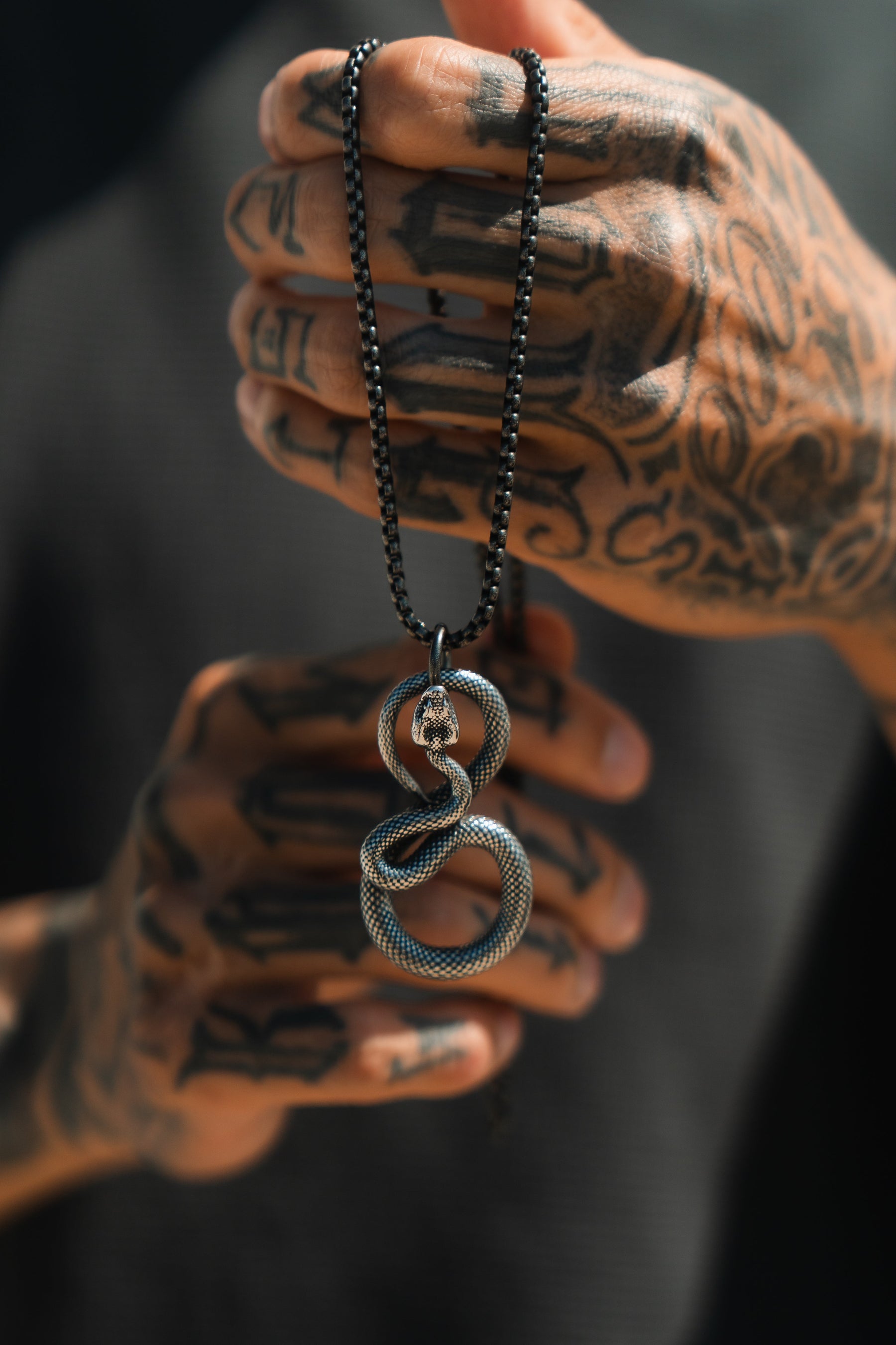 Tattooed hands display the Slither Necklace by Relentless Betrayal, a bold statement piece featuring an intricate snake pendant on a black beaded chain. The dark background highlights the jewelrys detail and complements the striking tattoos.