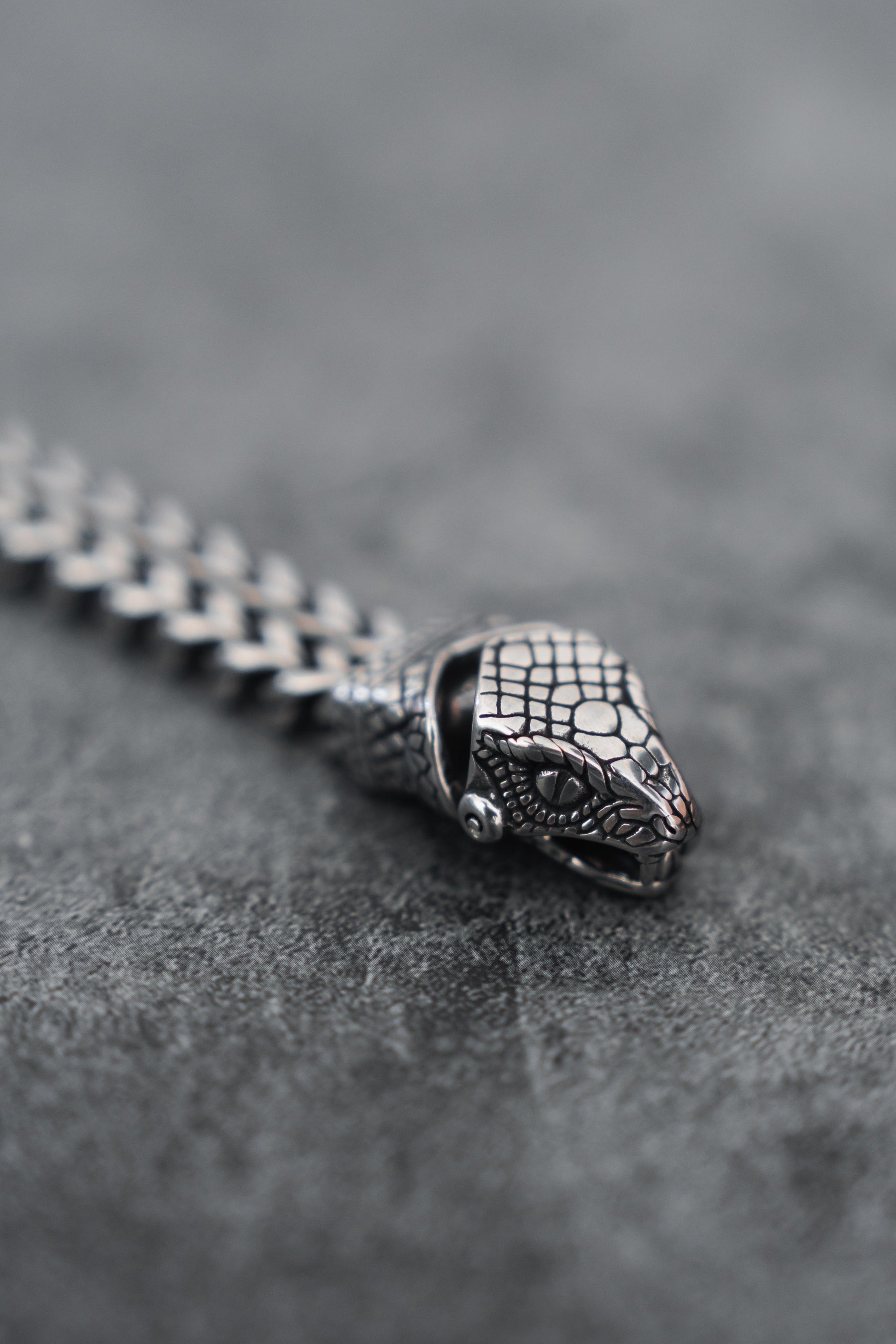 A close-up of the Relentless Betrayal Viper Bracelet in stainless steel, showcasing a snake head with detailed scales on a woven metal chain resting on a smooth, dark gray surface.