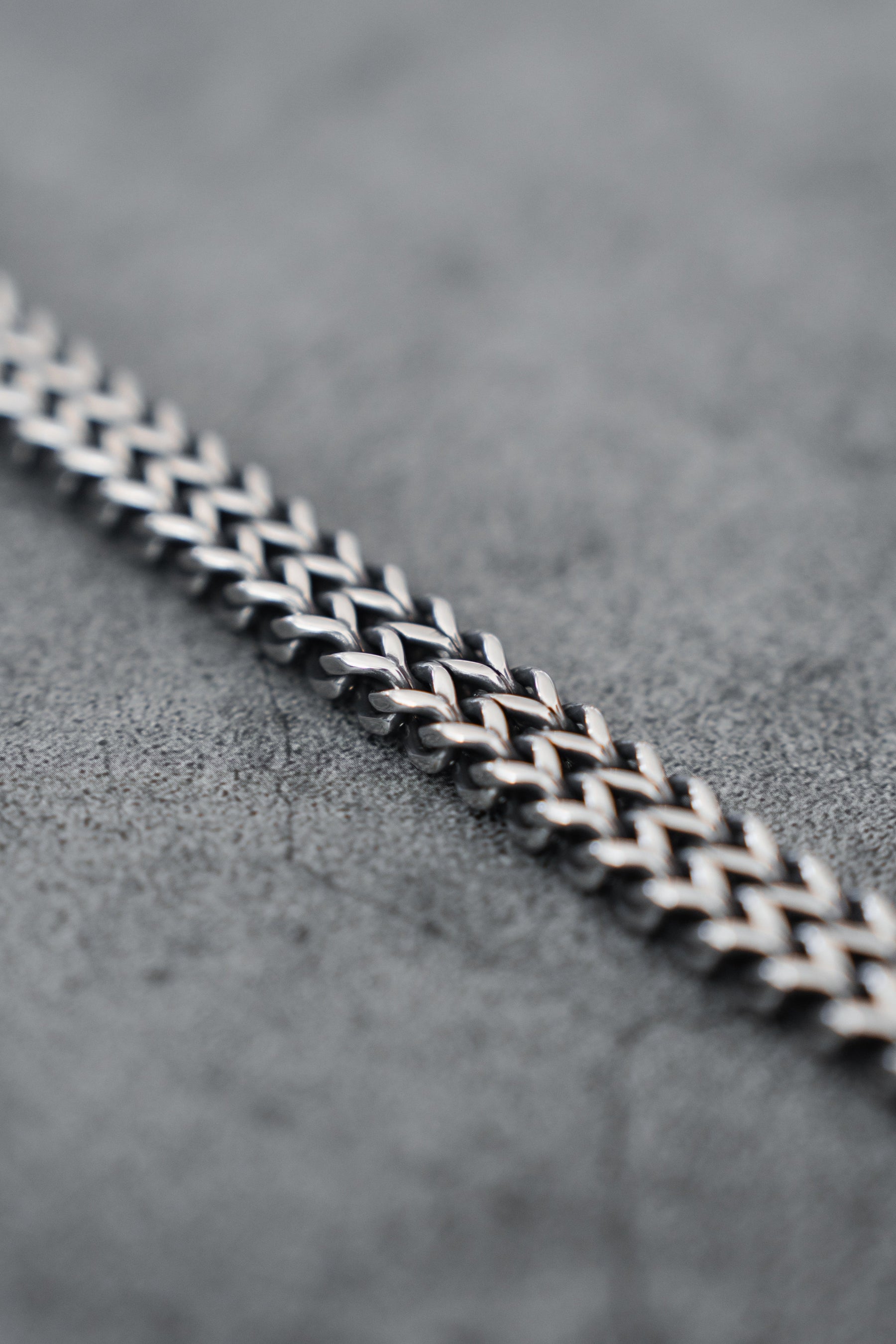 Close-up of the Relentless Betrayal Viper Bracelet in stainless steel, showcasing an intricate woven pattern on a gray textured surface. The bracelets reflective finish creates a striking contrast against the matte background.