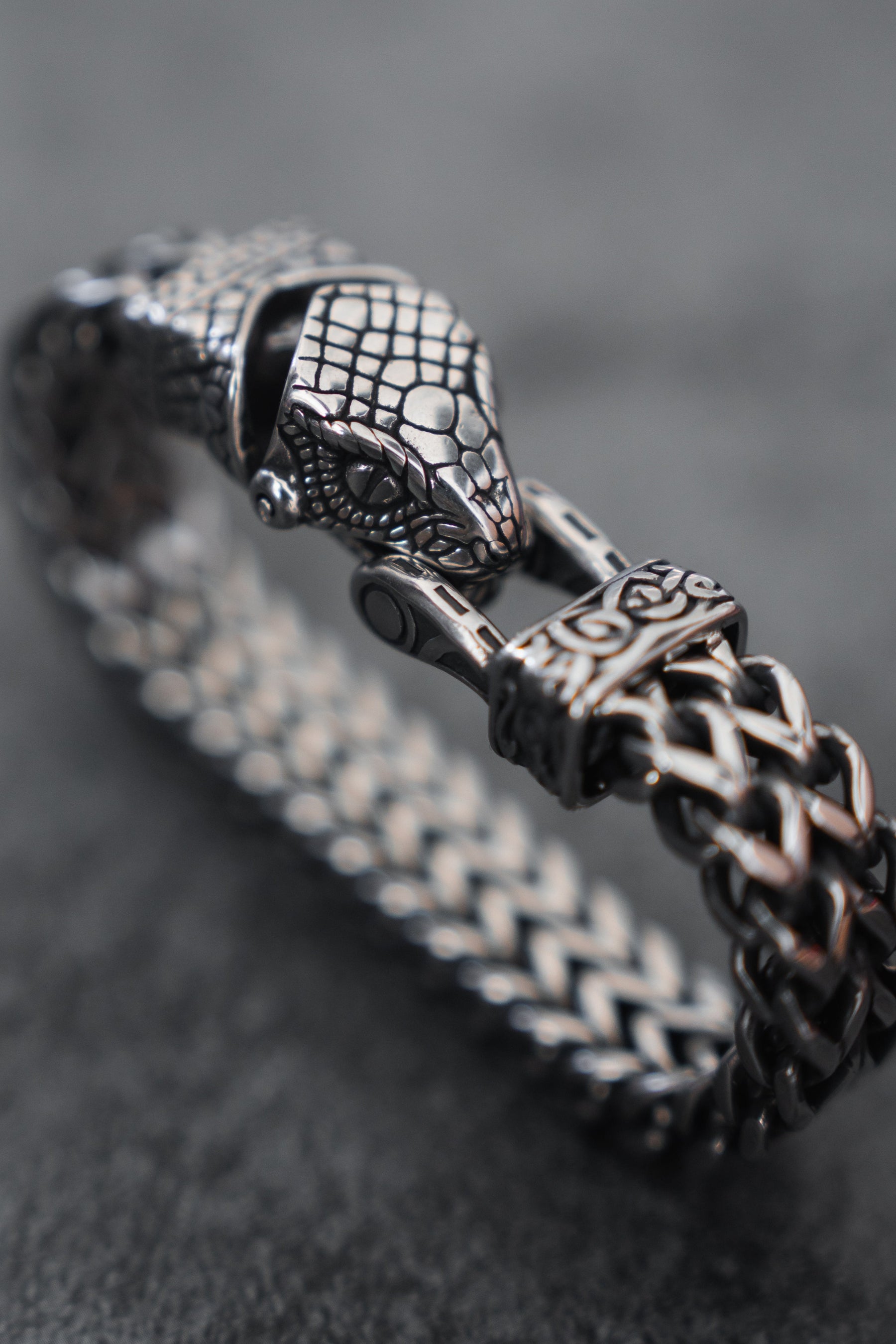 A close-up of the Viper Bracelet by Relentless Betrayal, crafted in stainless steel with a snake head design. This jewelry features a textured scale surface and woven chain pattern, set against a smooth, dark background.