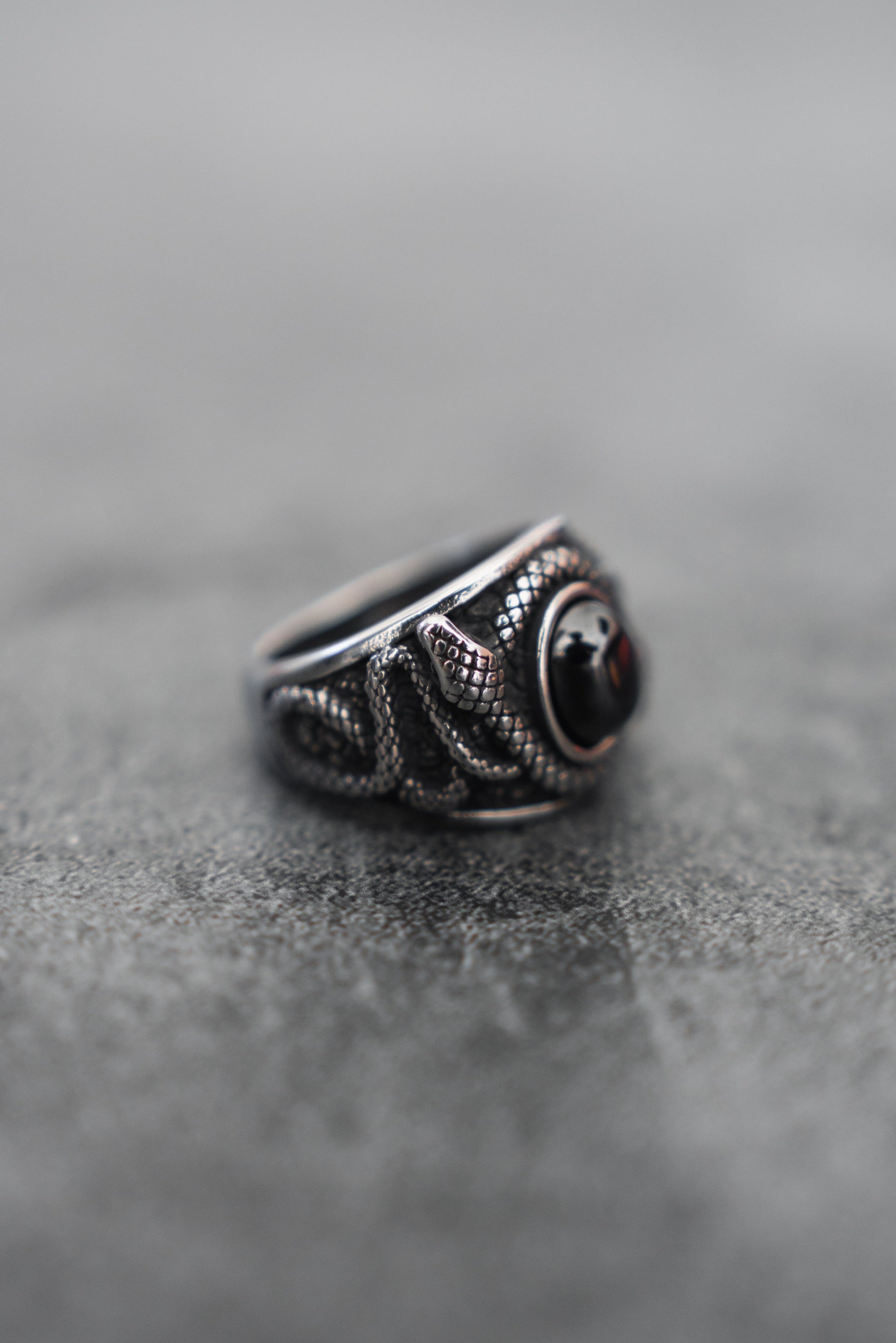 The Relentless Betrayal Onyx Rebirth Ring showcases detailed snake-like patterns around stainless steel, centered by a striking round black gemstone against a gray textured background.