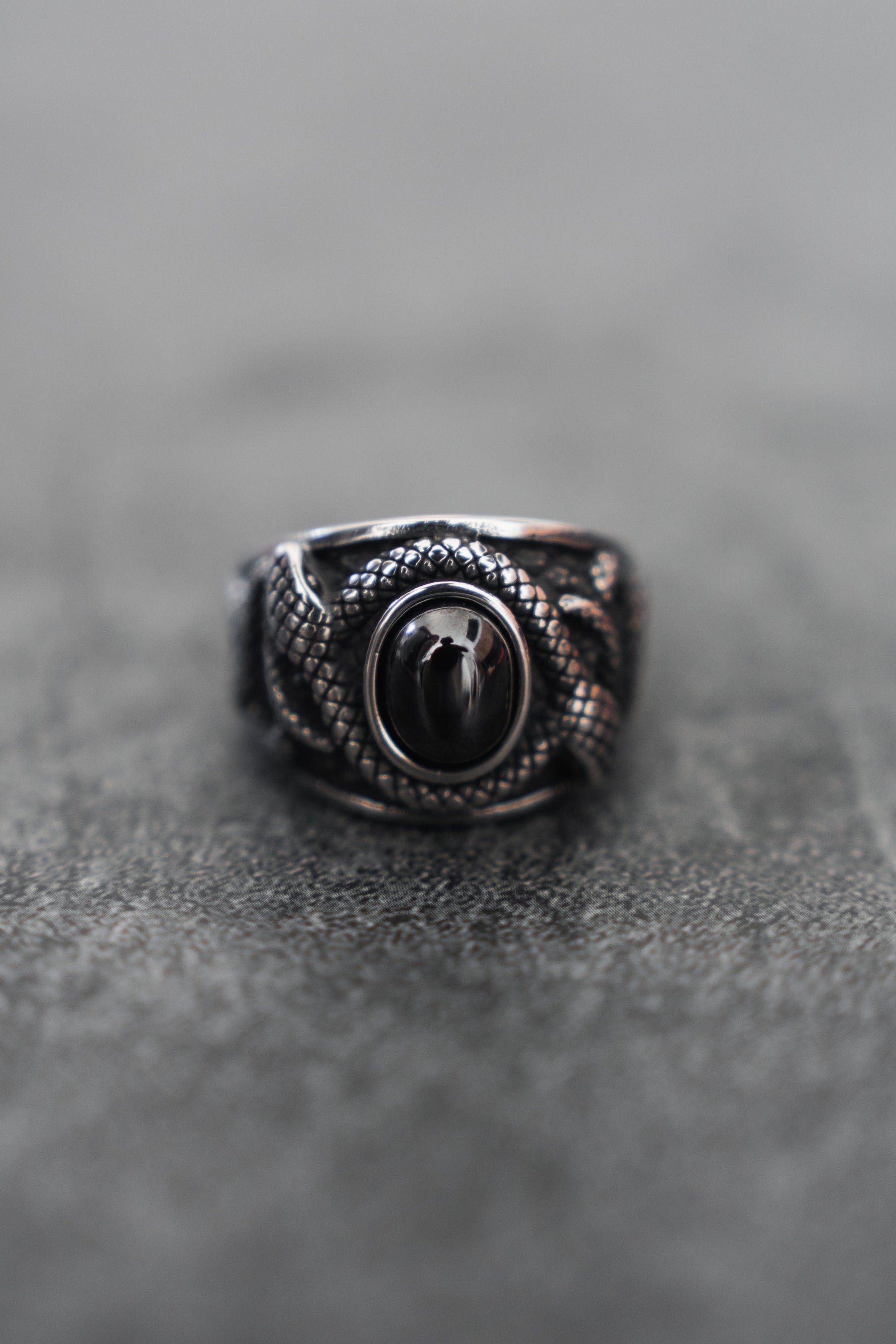 Discover the Onyx Rebirth Ring by Relentless Betrayal, a handcrafted silver ring with a snake design encircling a central black stone. Its intricately scaled serpentine form lies on a textured gray surface.