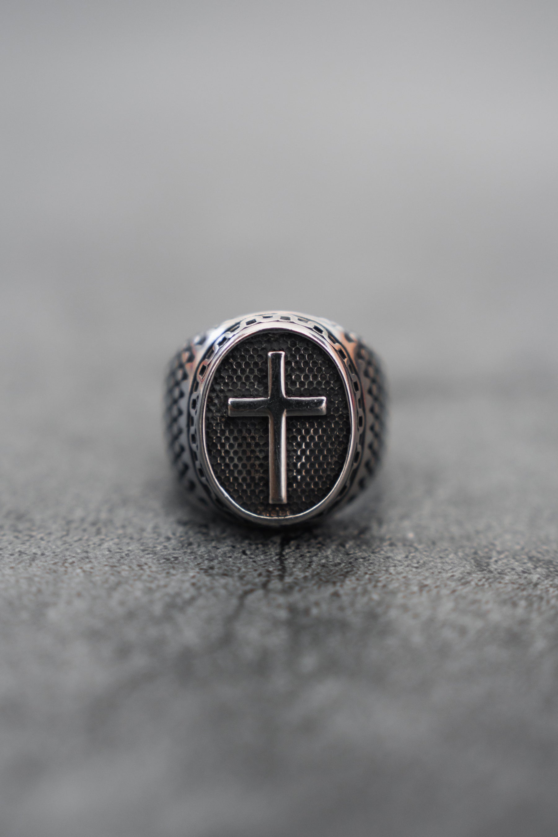 The Risen Savior Ring-Silver by Relentless Betrayal is a handcrafted stainless steel piece featuring an engraved cross on its textured oval face. The bands intricate details contrast against its gray, rough surface, enhanced by a blurred background.
