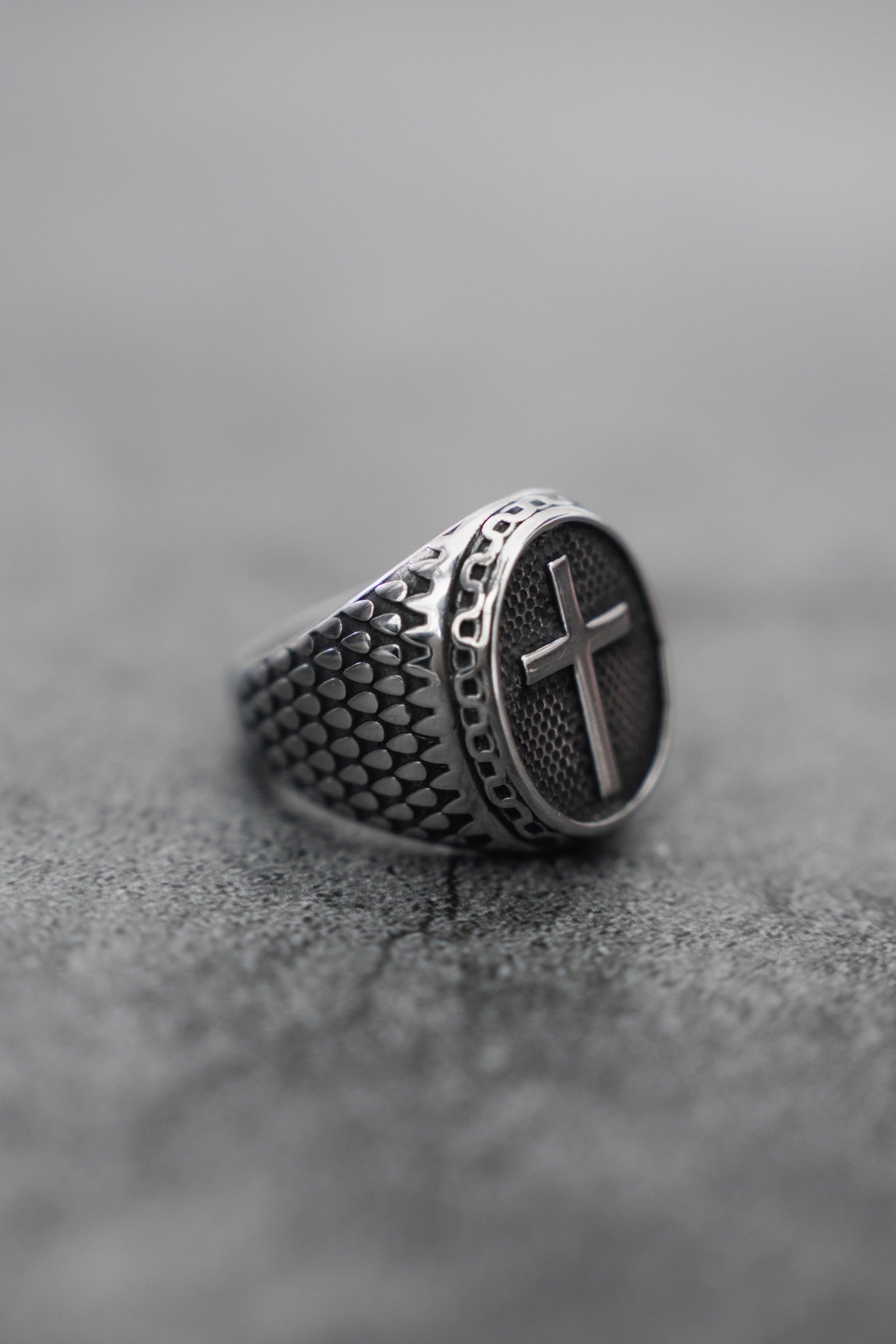 The Relentless Betrayal handcrafted Risen Savior Ring- Silver features a textured stainless steel band and a prominent cross on its face, elegantly displayed on a gray, slightly rough surface.