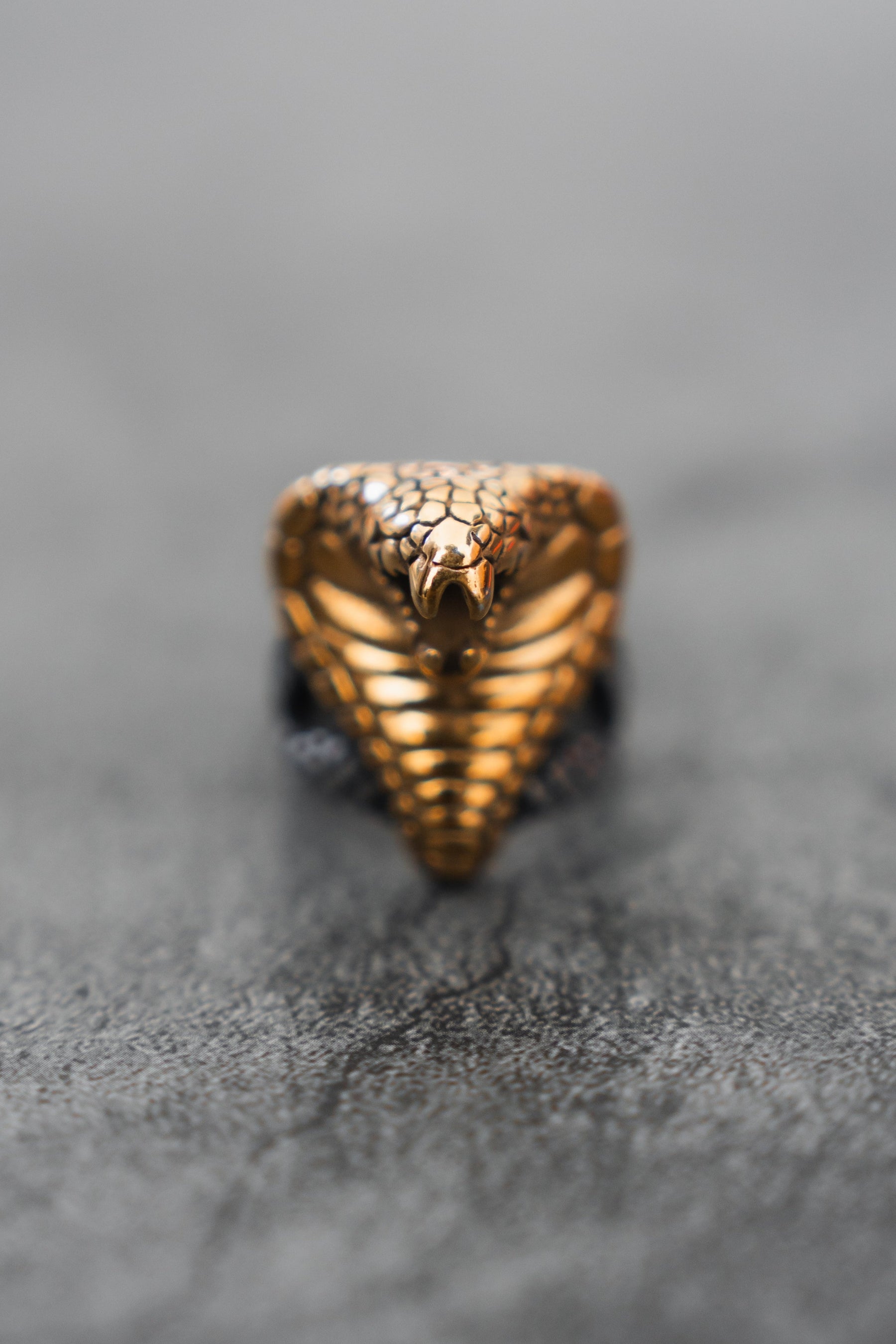 A detailed close-up of the Cobra King Ring by Relentless Betrayal shows its cobra-like design, intricate gold-colored stainless steel scale patterns, and a prominent open-mouthed head against a blurred gray background.