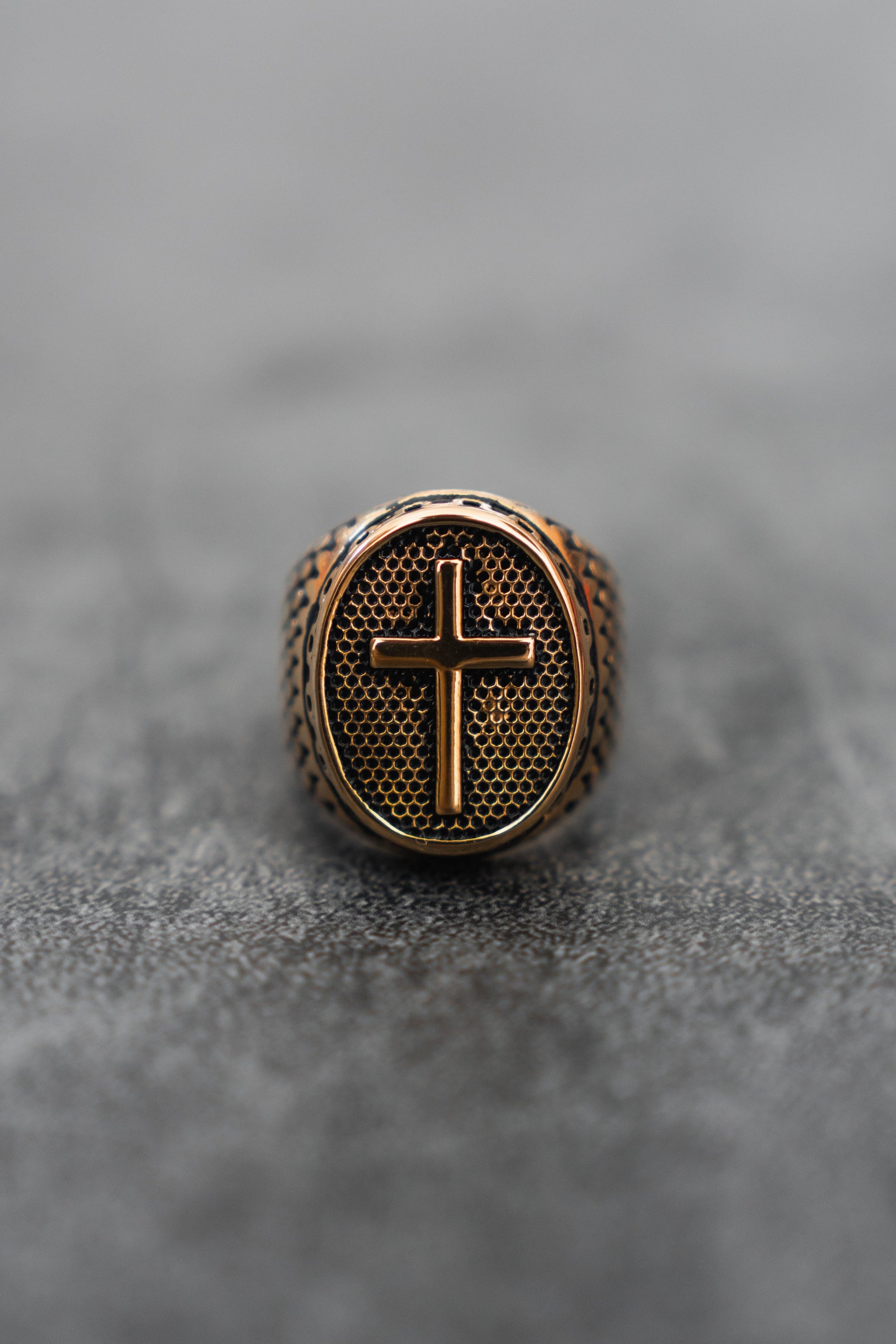 The Risen Savior Ring- Gold by Relentless Betrayal features a handcrafted bronze oval surface with a raised cross, set on a gray, blurred background.