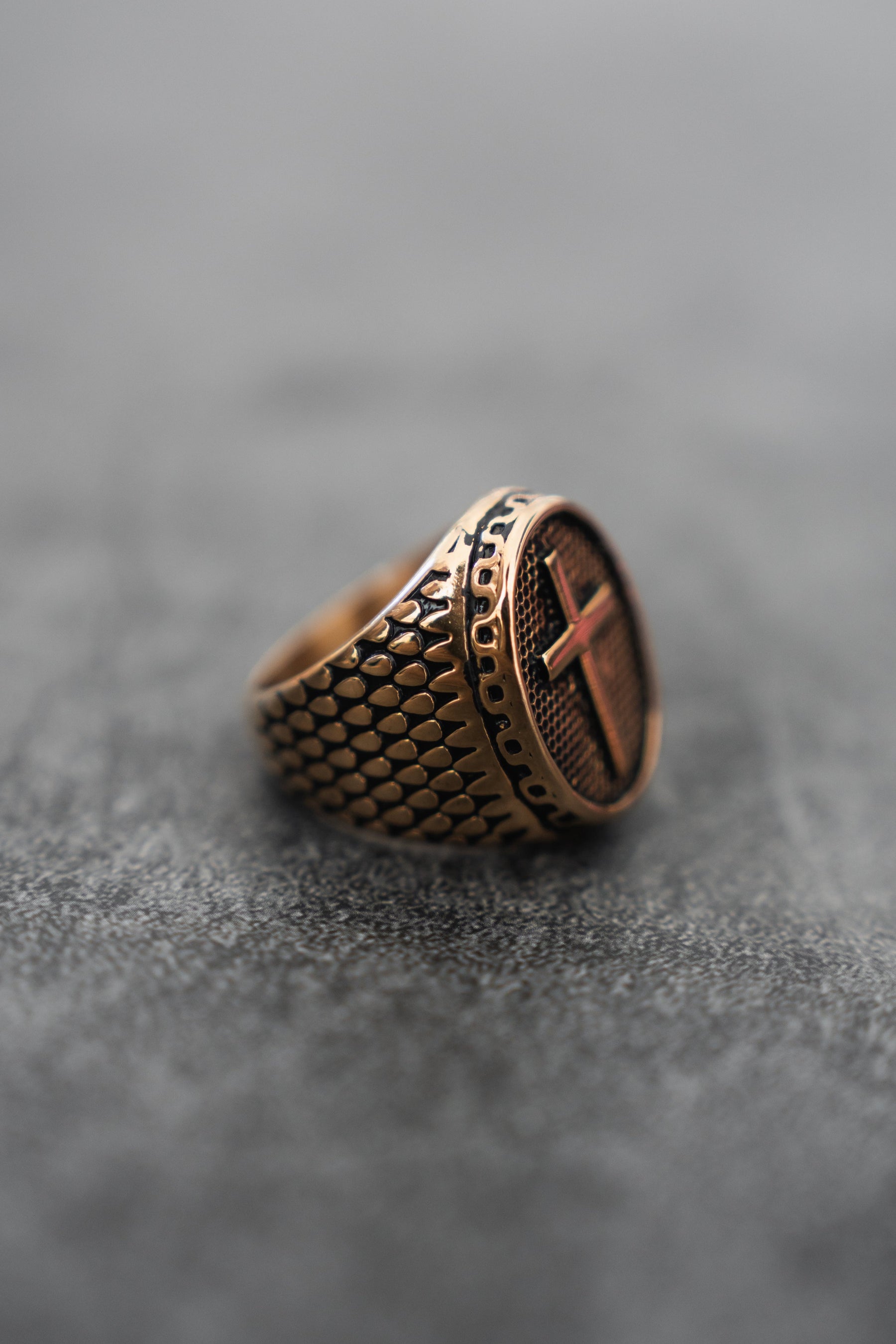 The Risen Savior Ring by Relentless Betrayal features a gold band with intricate textures and a circular face engraved with a cross design, resting gracefully on a textured gray surface.