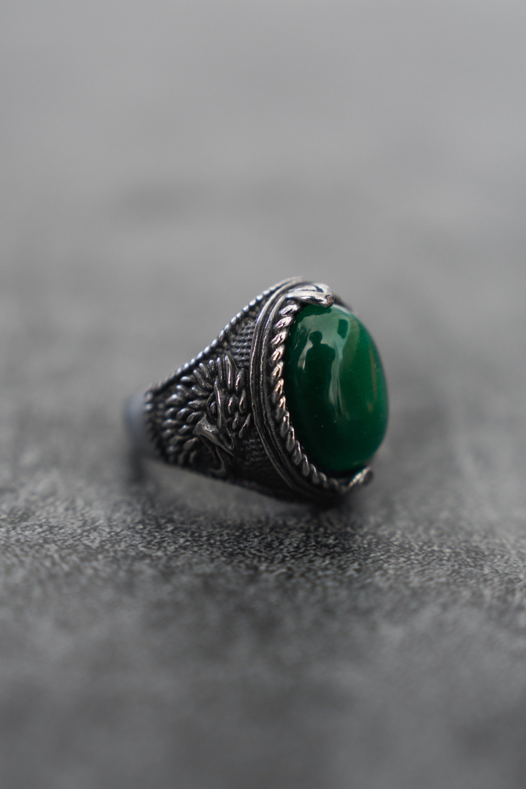 Relentless Betrayals Serpents Eye Ring, crafted in stainless steel, features intricate engravings and a central oval green gemstone that shimmers with an enchanting allure on a textured gray surface.