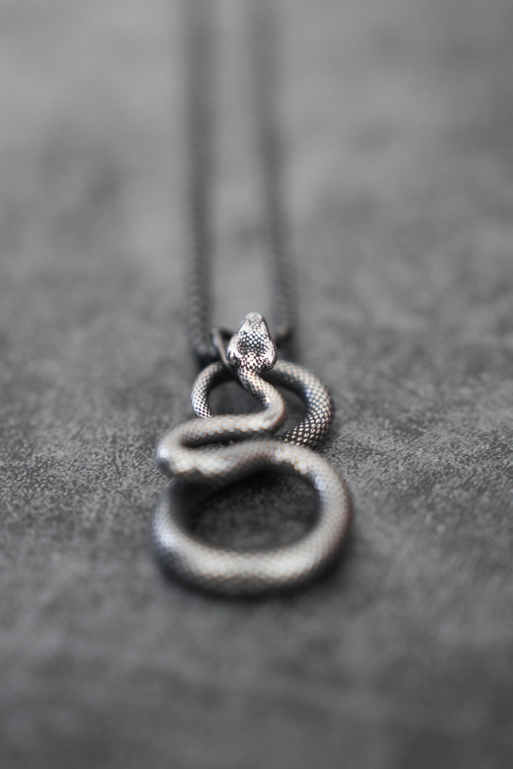 A silver Slither Necklace from Relentless Betrayal rests on a gray surface, showcasing intricate scales and detailing. This statement piece coils elegantly into a circle with its head slightly raised, embodying serpentine elegance.