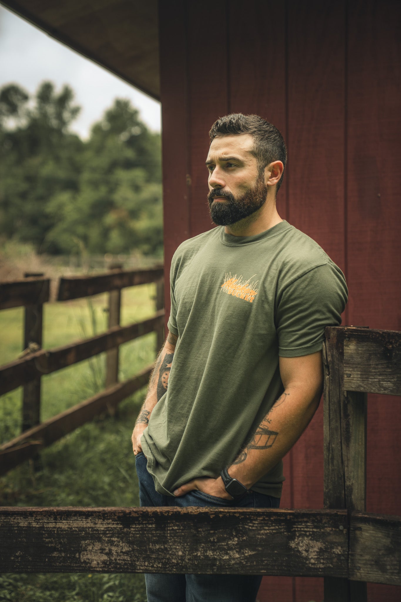 A bearded, tattooed man stands by a wooden fence outdoors, wearing Mucker Bucket’s Ghost Gobbler Premium Tee in olive green and jeans, with trees and a red barn in the background.