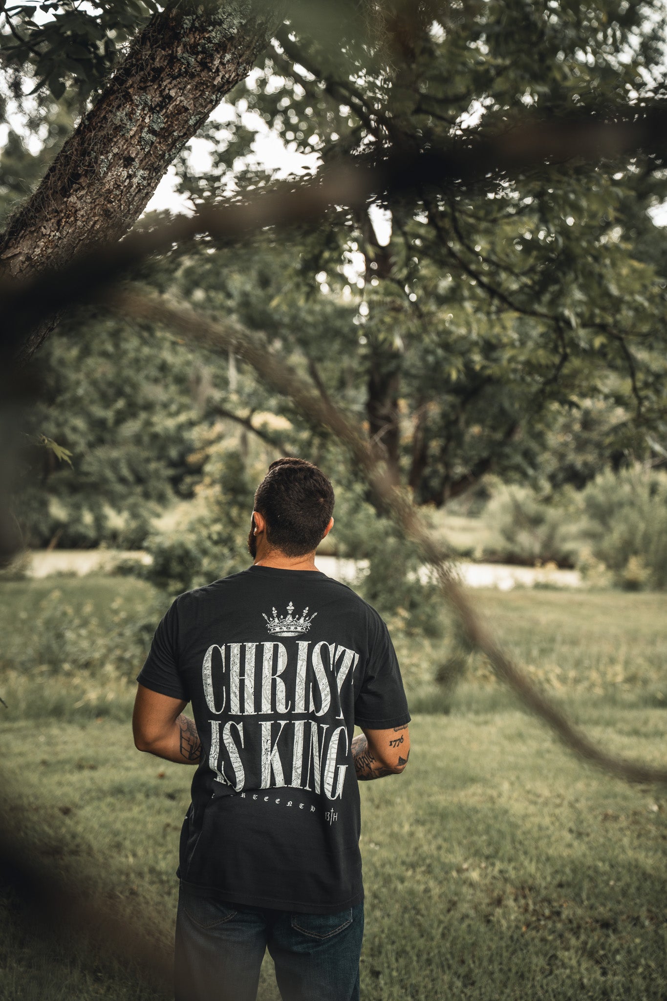 A man stands on grass near a tree, wearing the 13th brands Christ is King black T-shirt with bold white lettering on the back, set against a green, leafy background.