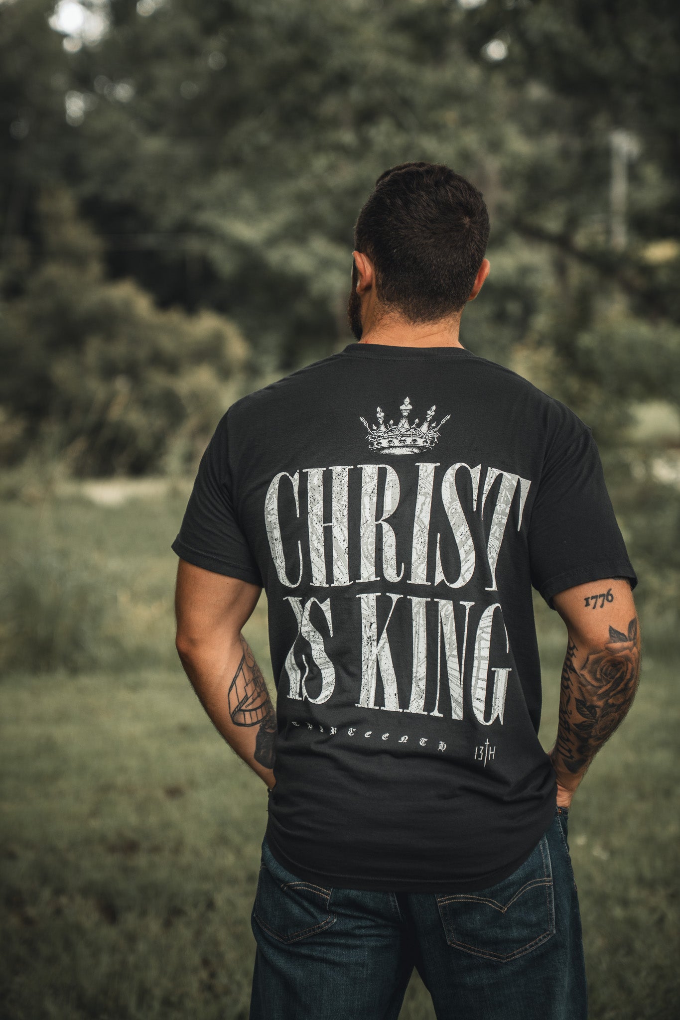 A man stands outdoors, back to the camera, wearing the 13th Christ is King black t-shirt with bold white crowned lettering. His tattooed arms are visible amidst green surroundings.