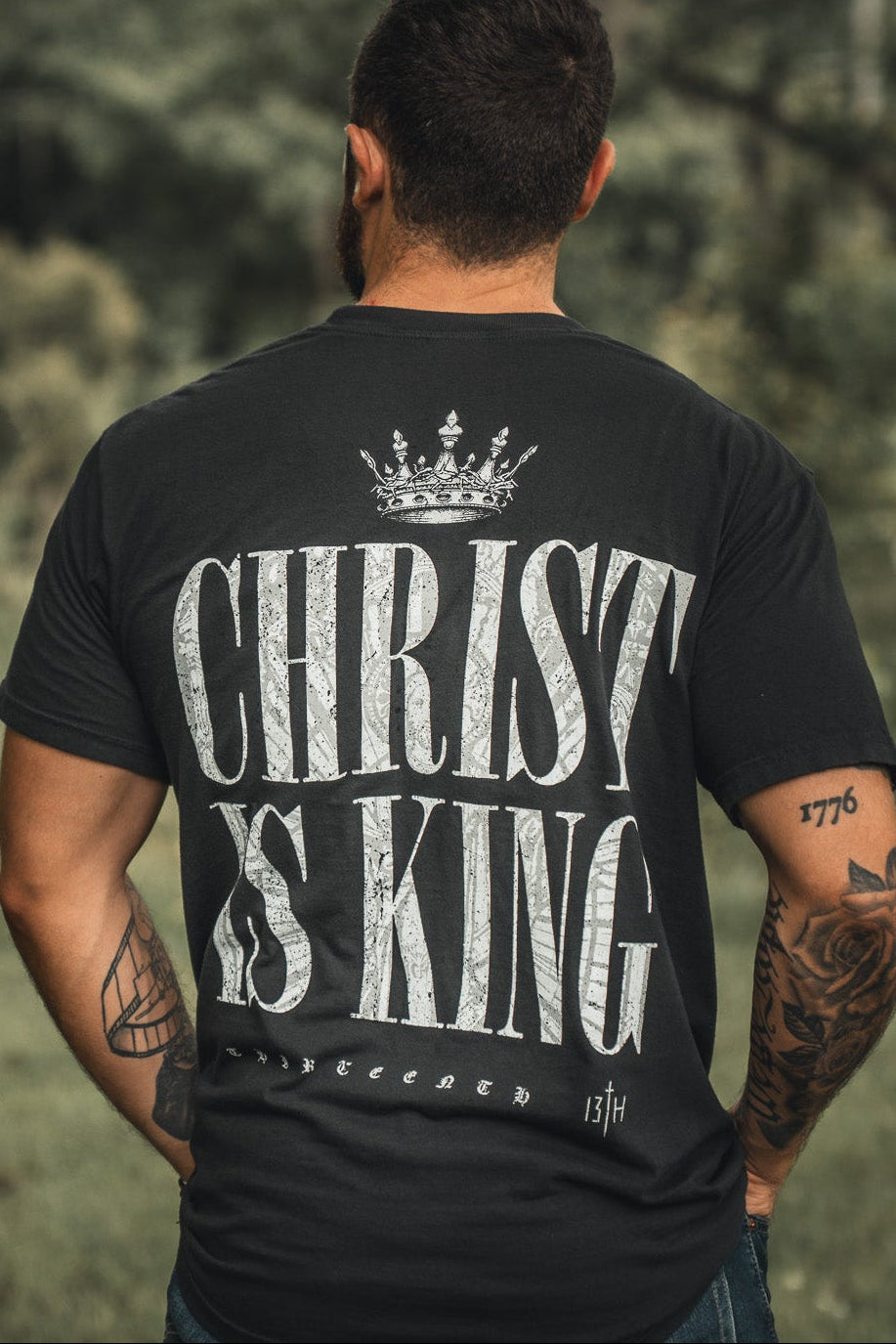 A man stands outdoors, back to the camera, wearing the 13th Christ is King black t-shirt with bold white crowned lettering. His tattooed arms are visible amidst green surroundings.