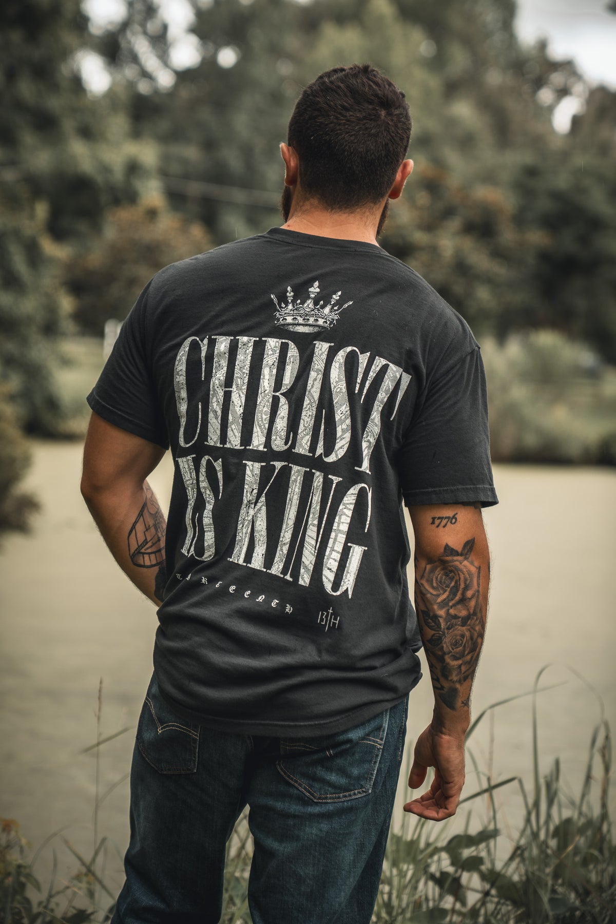 A tattooed man stands outdoors with his back to the camera, wearing the 13th brand Christ is King black T-shirt featuring bold white text and a crown graphic. Lush trees and greenery fill the background.