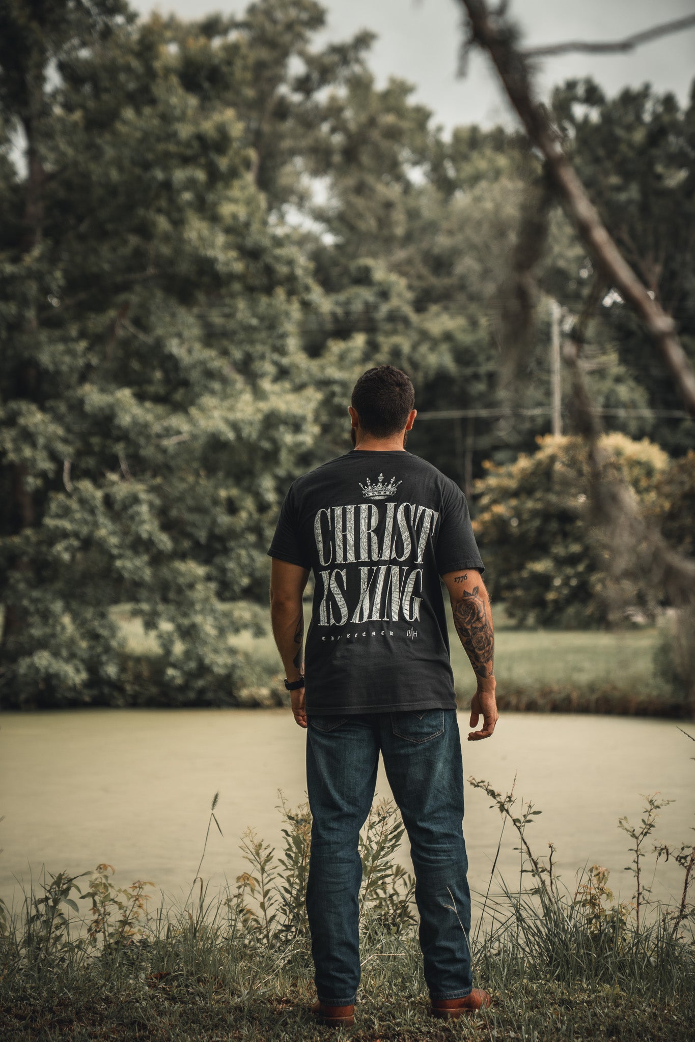 A man stands by a serene pond, wearing the 13th brand Christ is King black T-shirt with large white lettering, showcasing his faith as he faces away amid lush greenery.