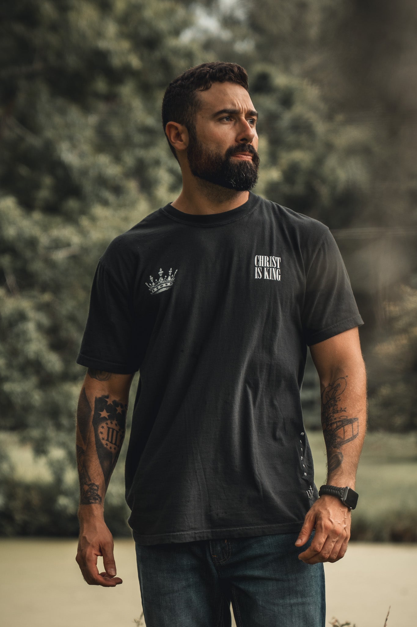 A bearded, tattooed man wearing a black Christ is King t-shirt by 13th and jeans stands outdoors before blurred green trees, looking to the side.