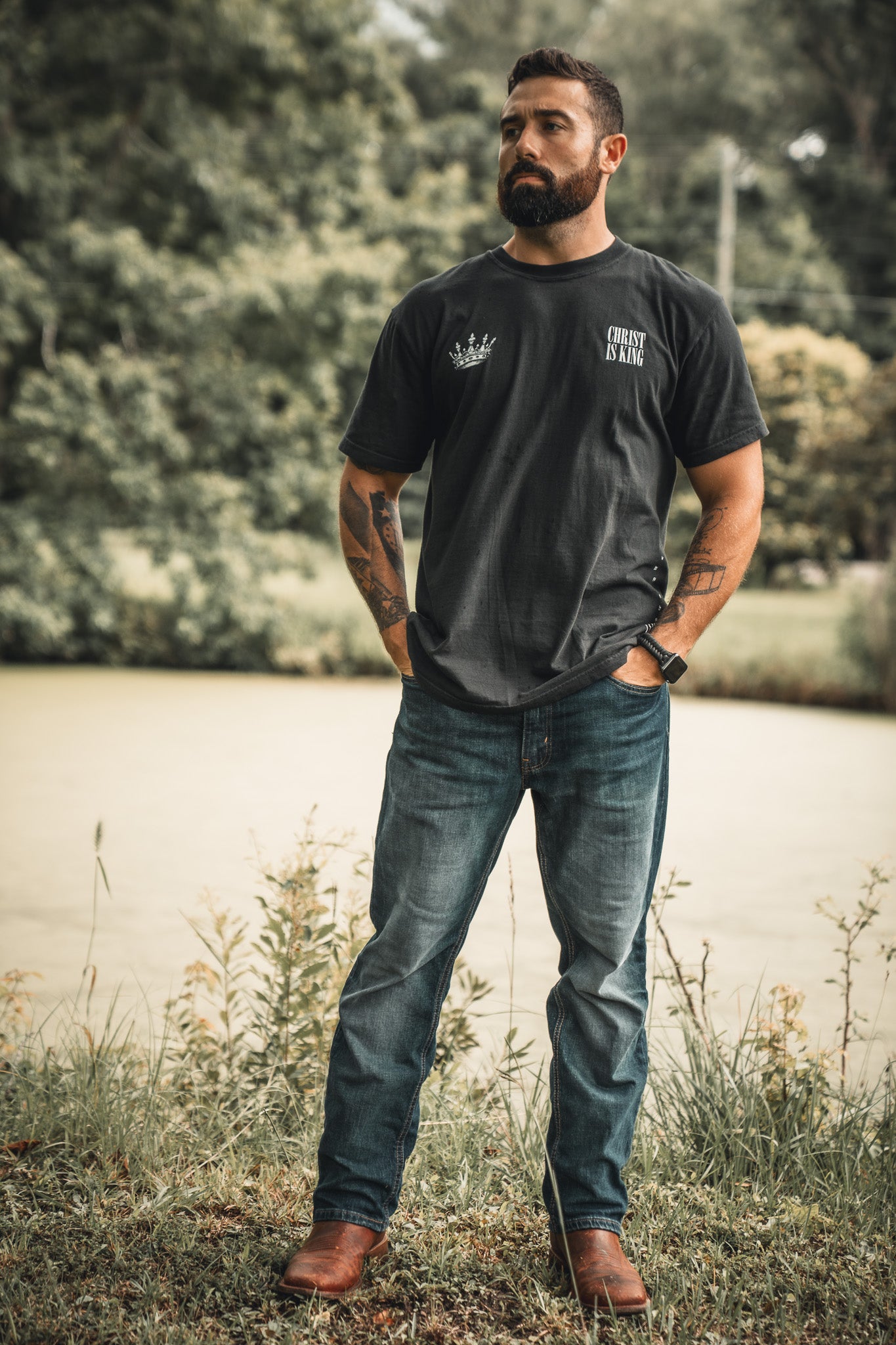 A man stands on grass near water, hands in pockets, wearing the 13th Christ is King black t-shirt, blue jeans, and brown boots. Amid trees and greenery, he gazes ahead with calm, quiet confidence.