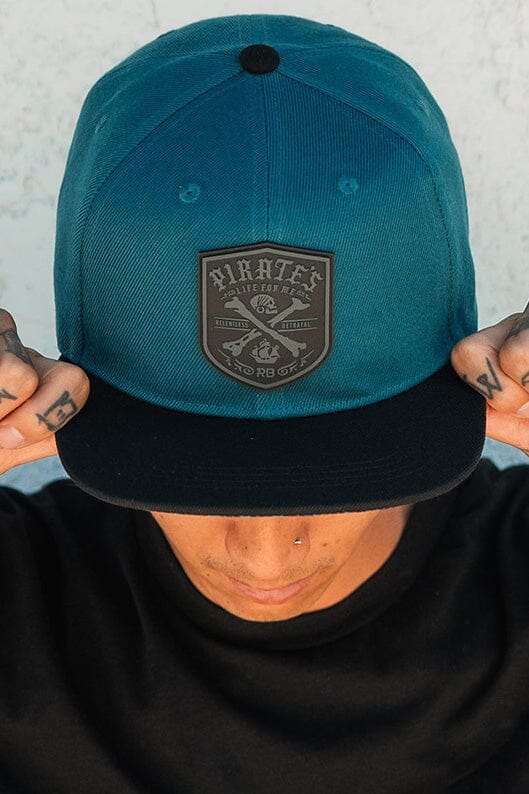 A person gazes down, wearing the Pirates Life Teal Snapback by Relentless Betrayal. Featuring PVC patches with a Pirates shield emblem and crossed swords, it complements their tattoos and pierced nose against a light backdrop, embracing the pirate lifestyle effortlessly.