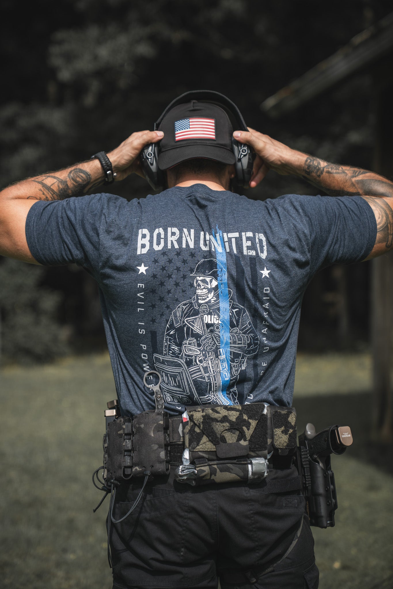 Standing outdoors, a person wears an American flag cap, headphones, tactical gear, and The Good Are Unafraid t-shirt by Born United Private Label featuring a police officer and flag graphic to show support for the Blue.