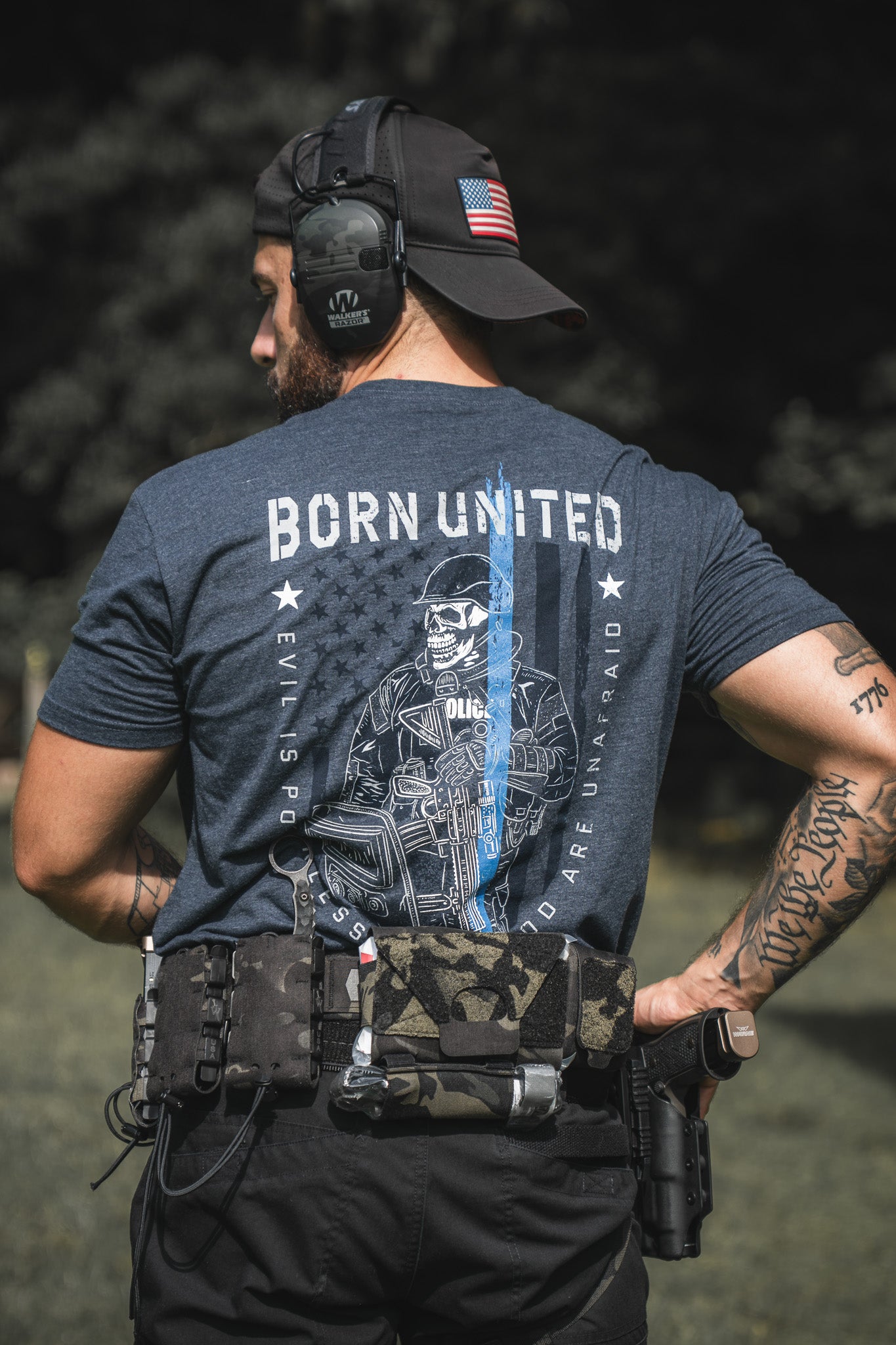 A man outdoors with tactical gear and a holstered firearm wears a backward US flag cap, ear protection, and Born United Private Labels The Good Are Unafraid t-shirt, showing his support for the blue.