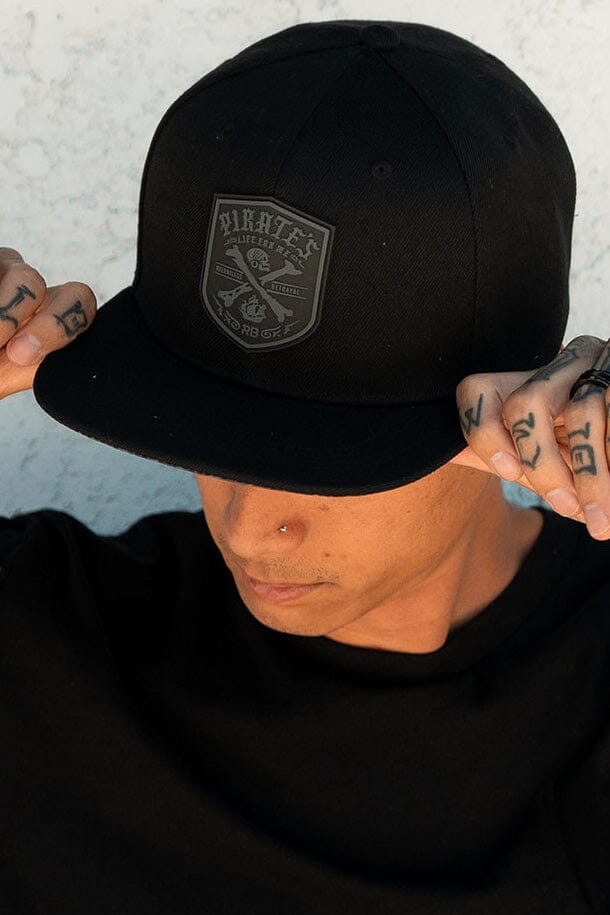 A person wearing the Pirates Life Black Snapback by Relentless Betrayal adjusts the brim with tattooed fingers, displaying a nose piercing against a textured white wall.