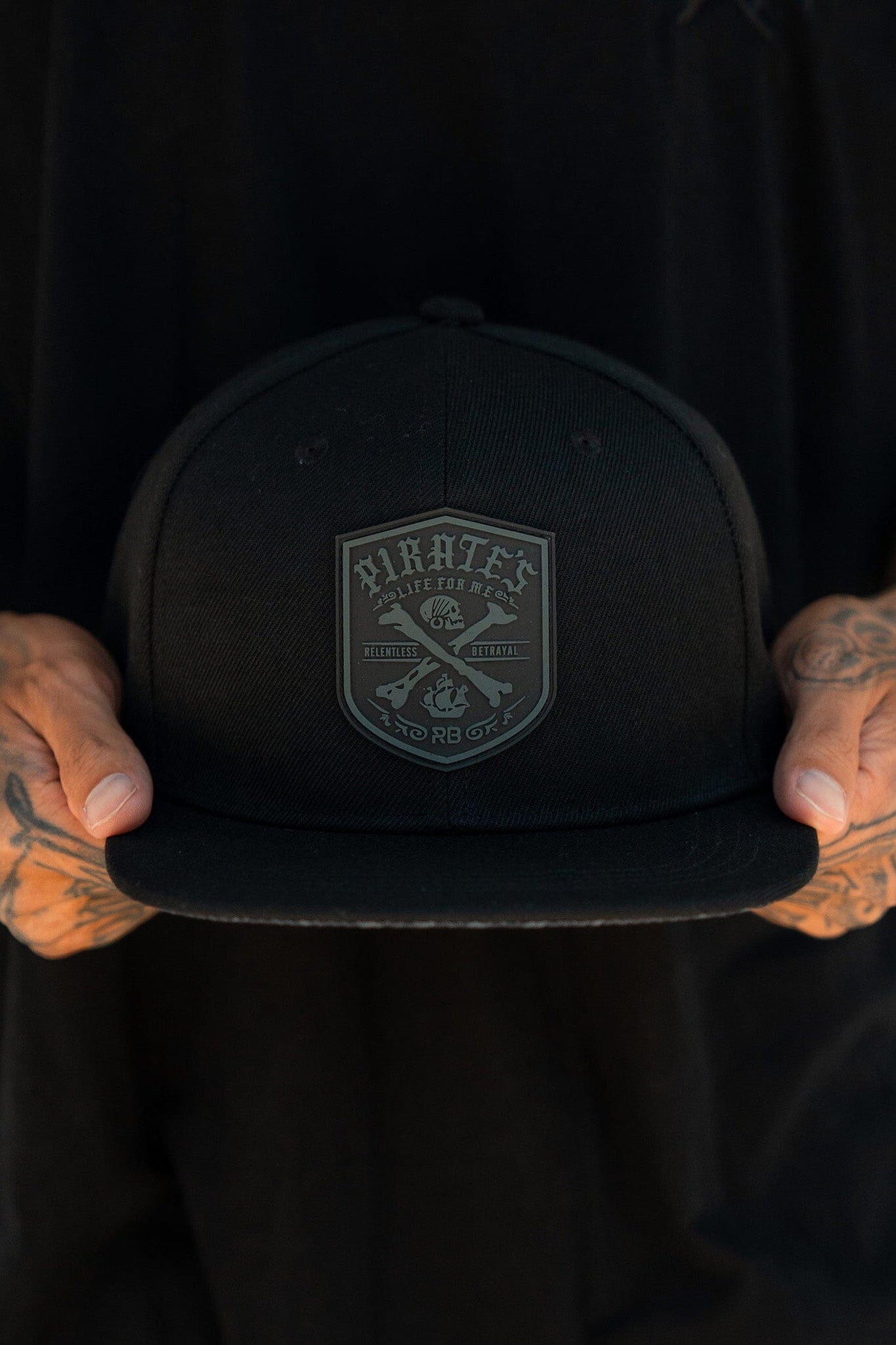 Amid a moody, dimly lit scene, a person holds the Relentless Betrayals Pirates Life Black Snapback. The cap features a custom PVC rubber patch with Pirates and intricate designs. Their dark shirt and tattooed hands complement the snapbacks secure fit.