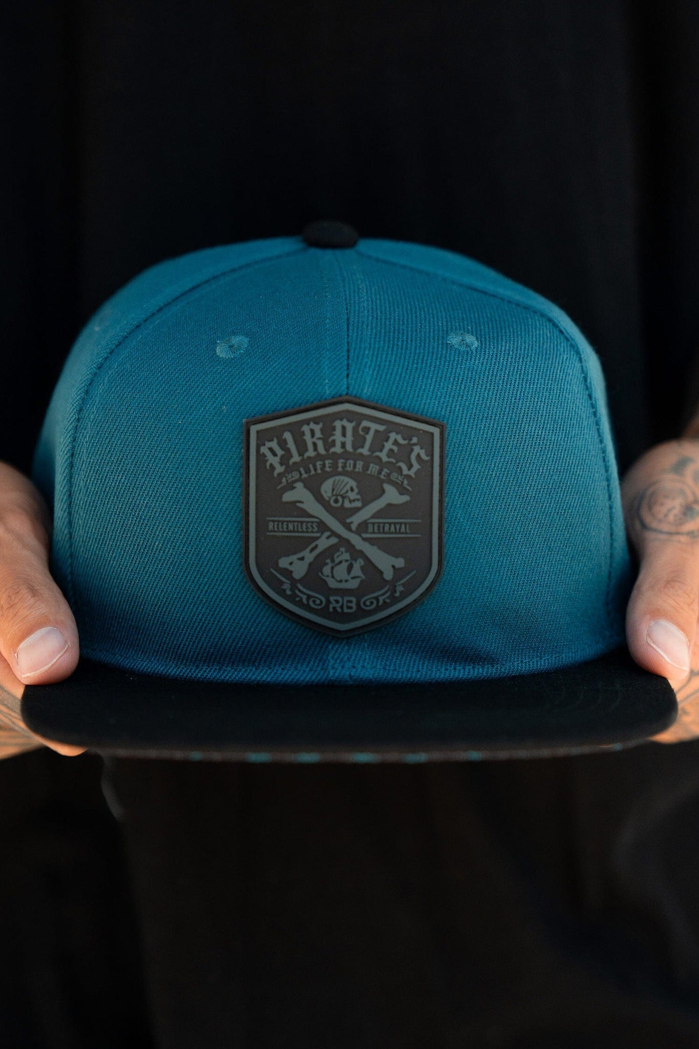 Someone holds the Pirates Life Teal Snapback by Relentless Betrayal, featuring a snapback enclosure and a detailed emblem reading Pirates Life for Me, surrounded by nautical symbols.