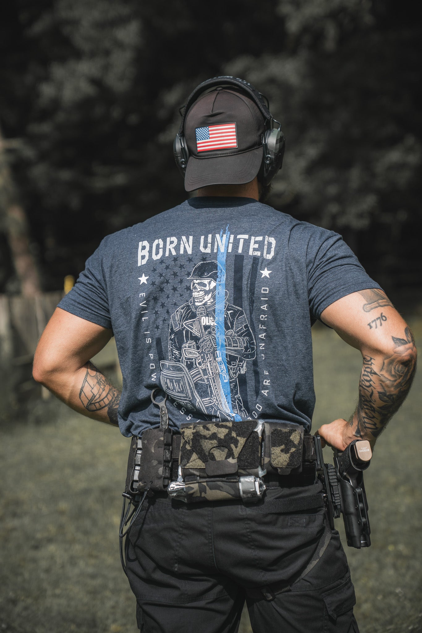 A person stands outdoors, hands on hips, wearing The Good Are Unafraid shirt by Born United Private Label with a police support graphic, plus a U.S. flag cap, headphones, and a utility belt holding gear and a firearm.