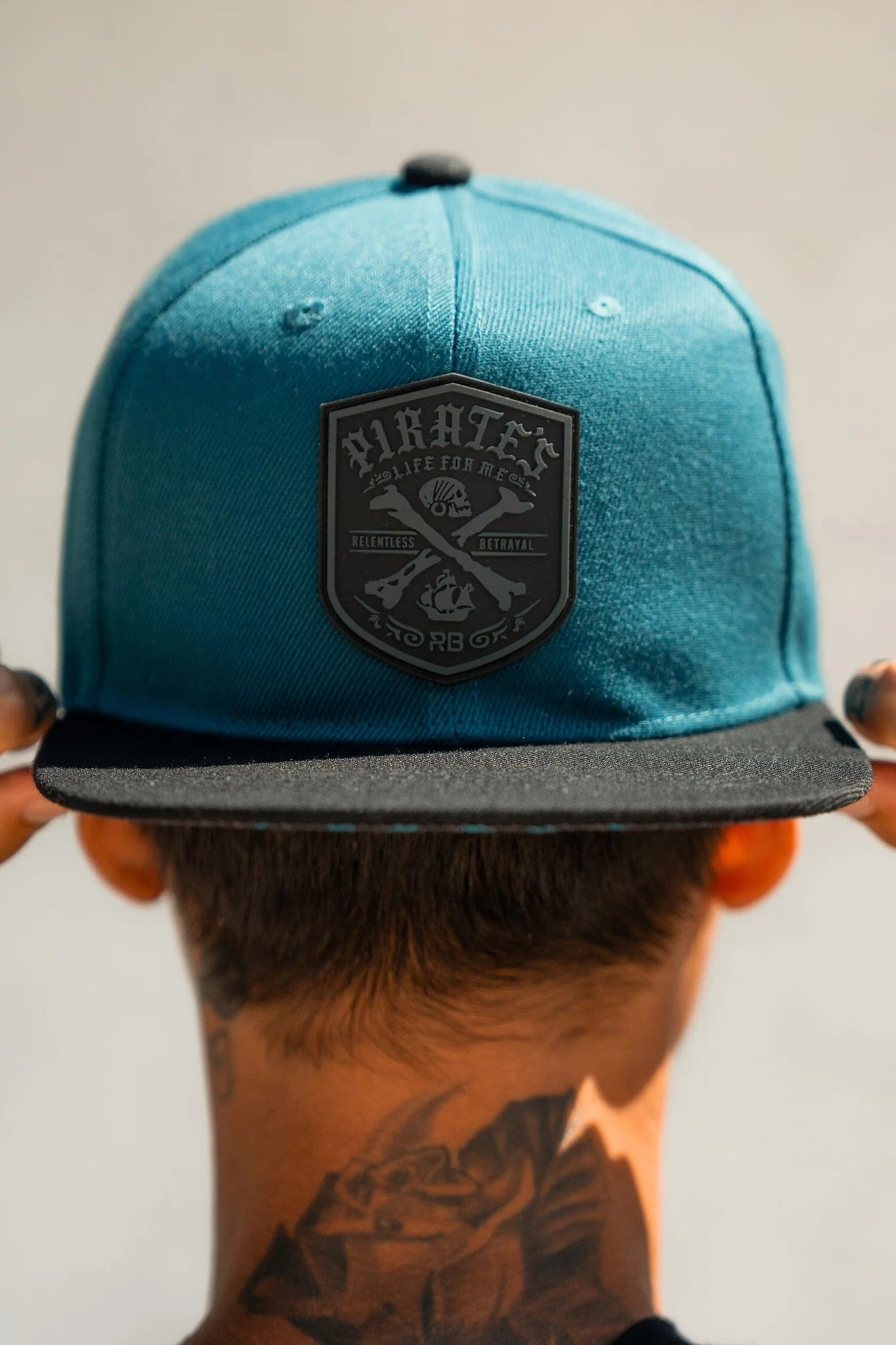 A neck-tattooed person sports the Relentless Betrayal Pirates Life Teal Snapback, showcasing a dark pirate emblem. Its black visor complements the sunny glow, and the snapback enclosure ensures a secure fit, embodying that adventurous lifestyle.