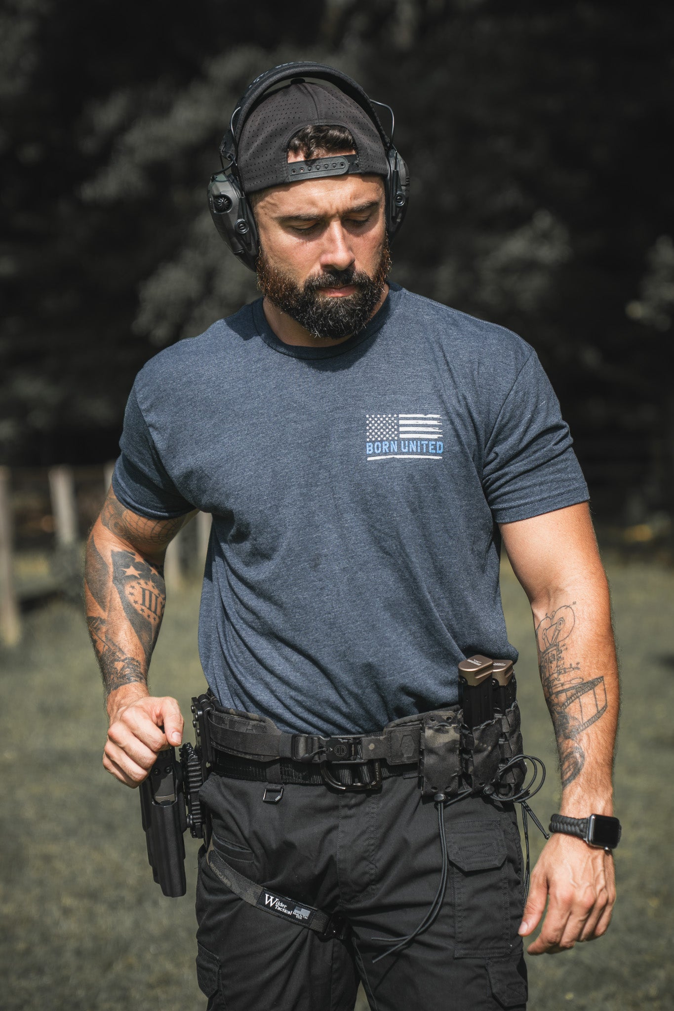 A bearded, tattooed man in black tactical pants and a backward cap wears The Good Are Unafraid T-shirt by Born United Private Label. Outdoors, with headphones and smartwatch, he holds a holstered gun and looks serious.