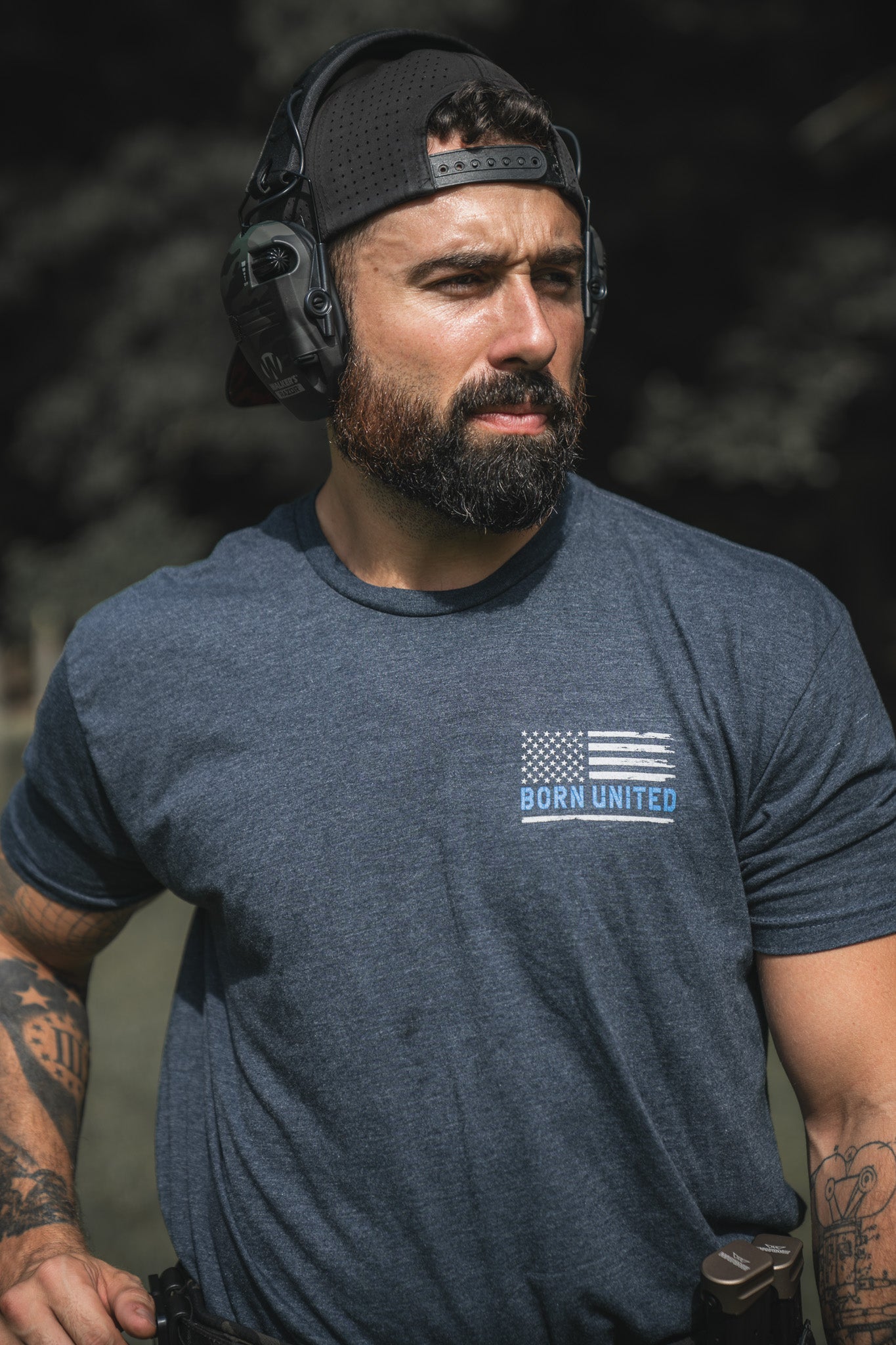 A bearded man in a backward cap and headphones wears a blue The Good Are Unafraid T-shirt from Born United Private Label outdoors, showing tattoos and a holstered gun at his waist, standing strong with a serious expression.